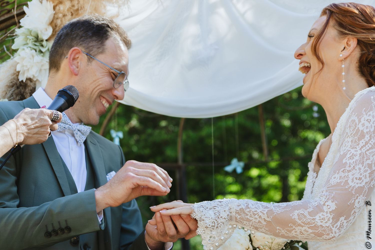 Groom laughs while placing ring on bride's finger during a joyful outdoor wedding ceremony exchange.