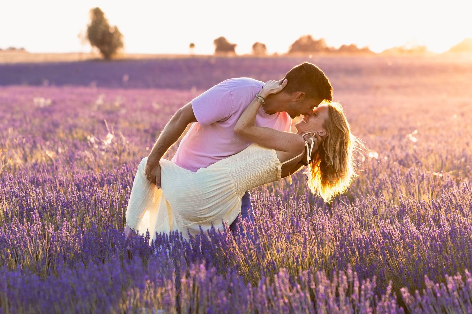 Romantic couple sharing a passionate dip kiss in a stunning purple lavender field at golden sunset.