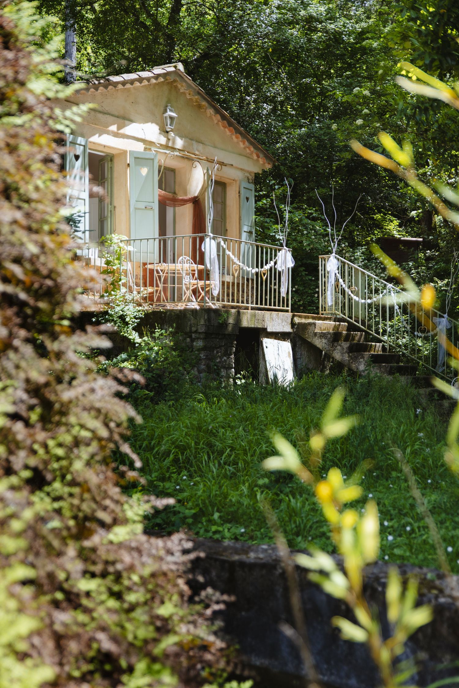 Charming cottage with green shutters, elevated deck, and lush garden surroundings in dappled sunlight.