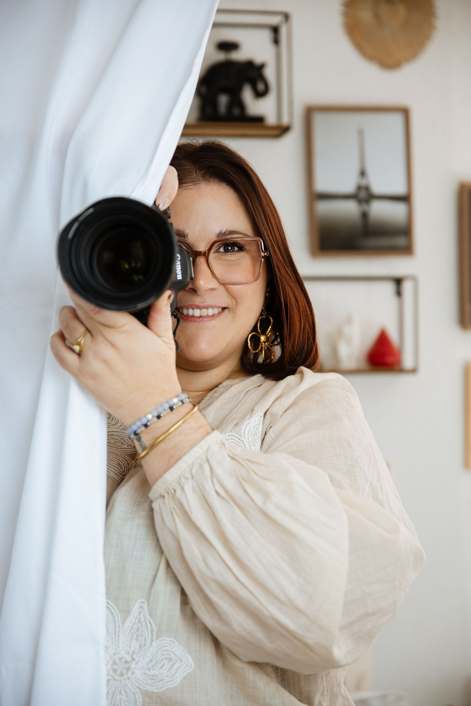 Smiling woman with glasses holding a professional camera, wearing a cream blouse and gold earrings, peeking around a white curtain.