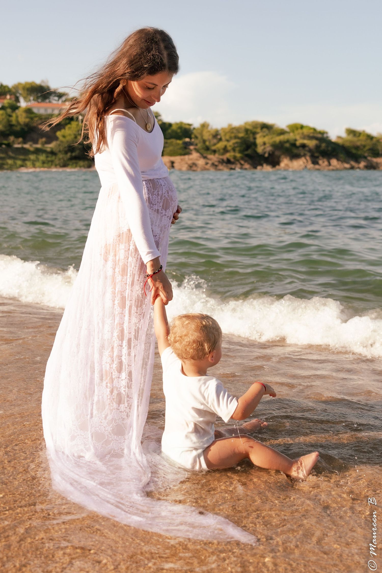 Pregnant woman in white lace dress holding toddler's hand at the beach during golden hour.