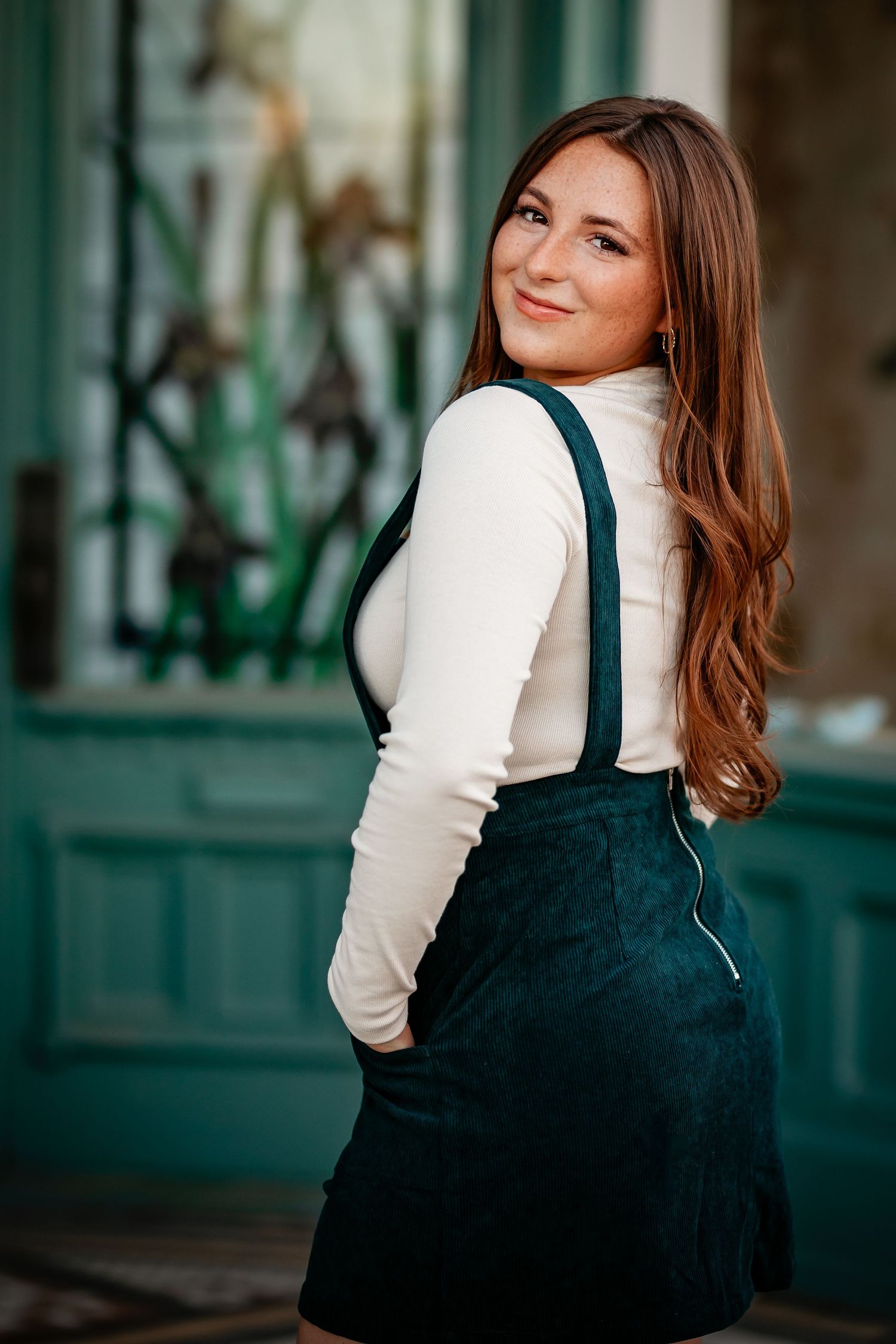 Brunette woman in green pinafore dress smiling over shoulder near teal door.