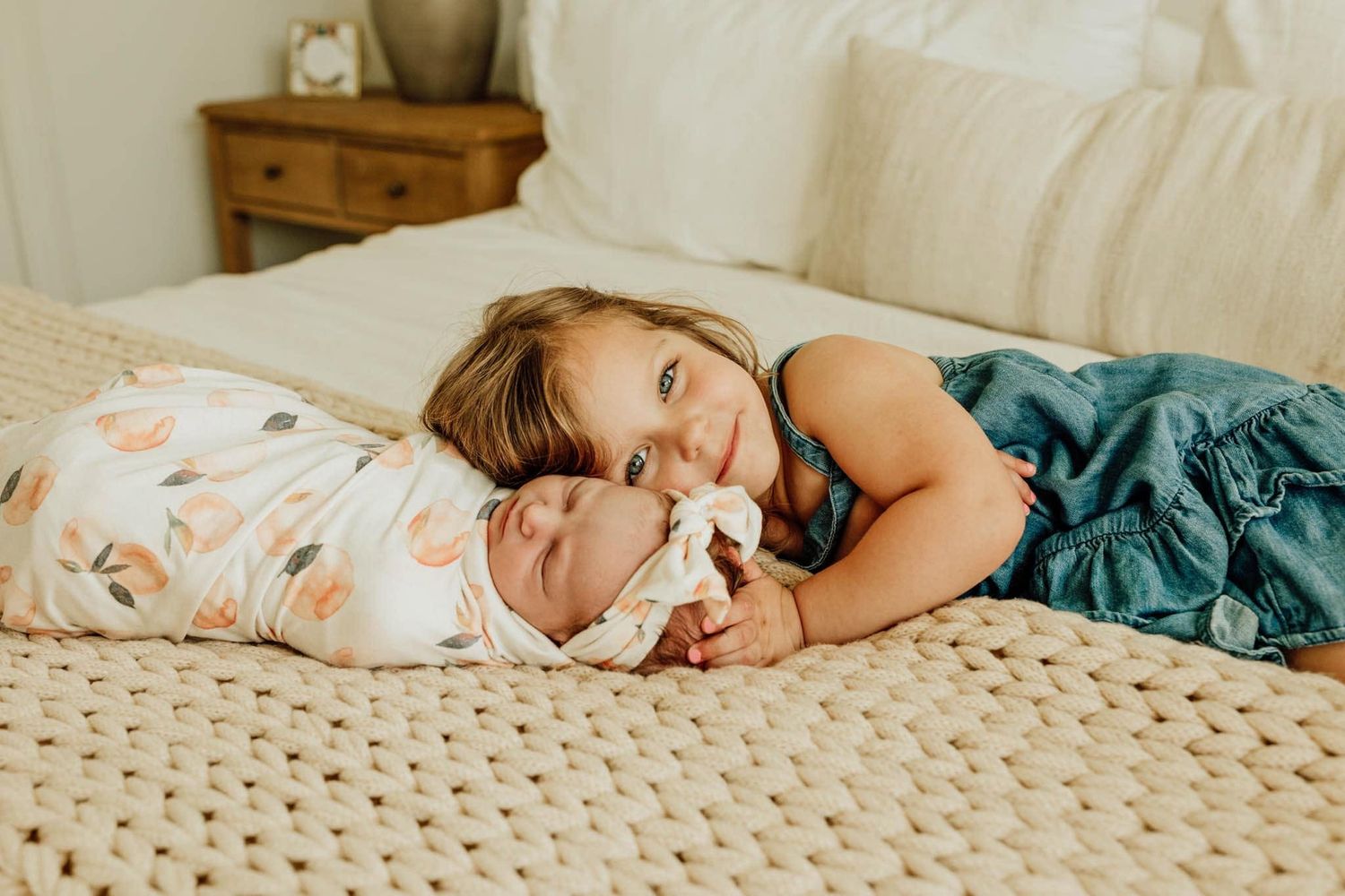 Young girl lying on bed cheek-to-cheek with swaddled newborn baby sibling in soft natural light.