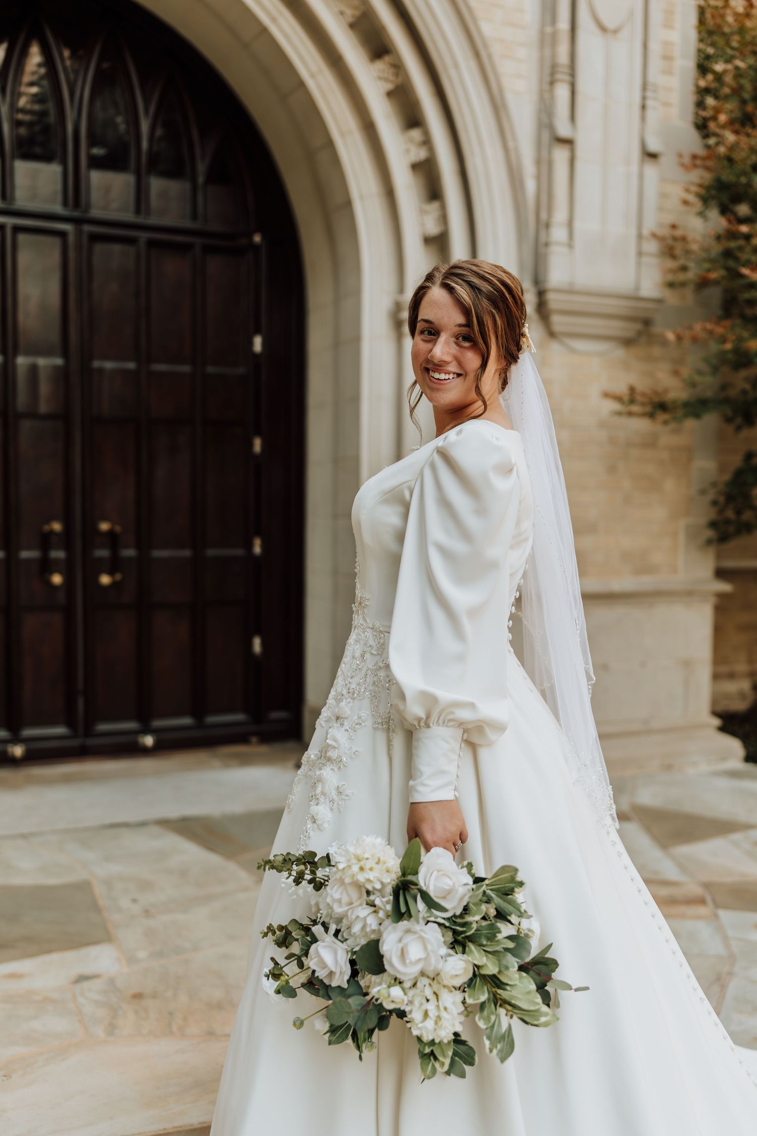 Elegant bride in long-sleeve white ball gown holding white bouquet outside stone church entrance.