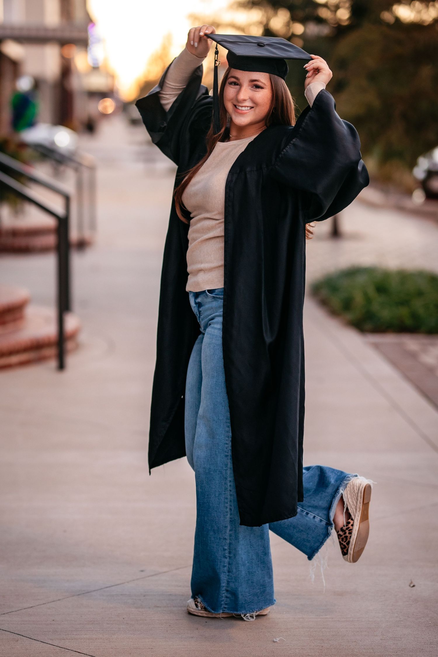 Smiling woman in black graduation gown and jeans posing on urban sidewalk.