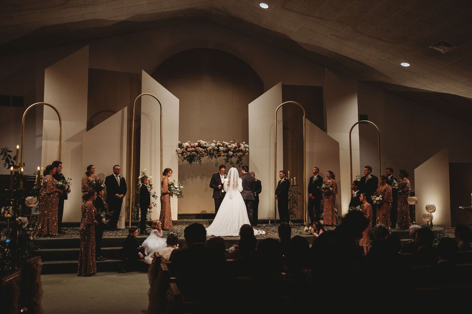 Bride and groom stand at altar during indoor church wedding ceremony with wedding party and candlelit decor.