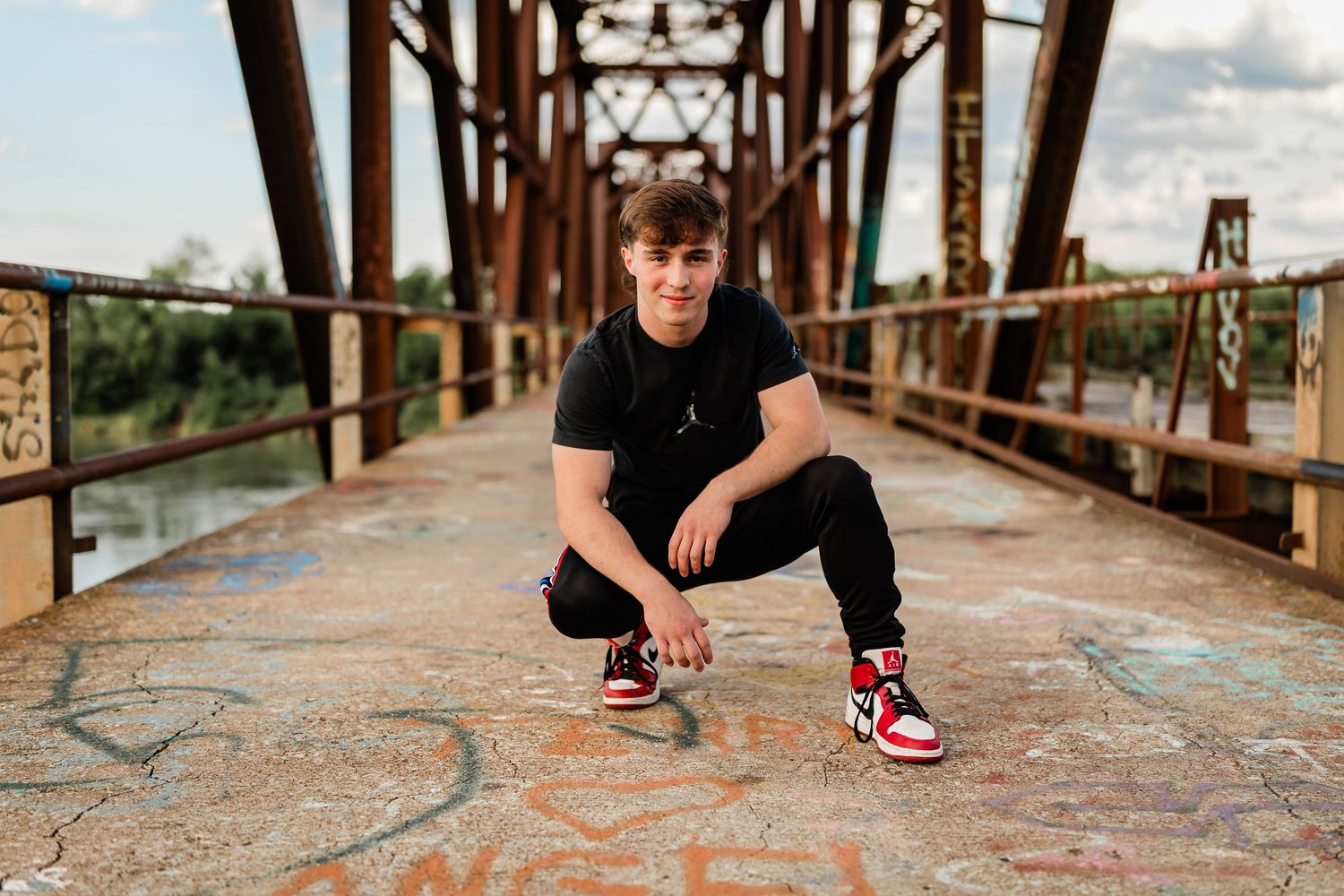 Young man in black outfit and Jordan sneakers crouching on a graffiti-covered bridge.
