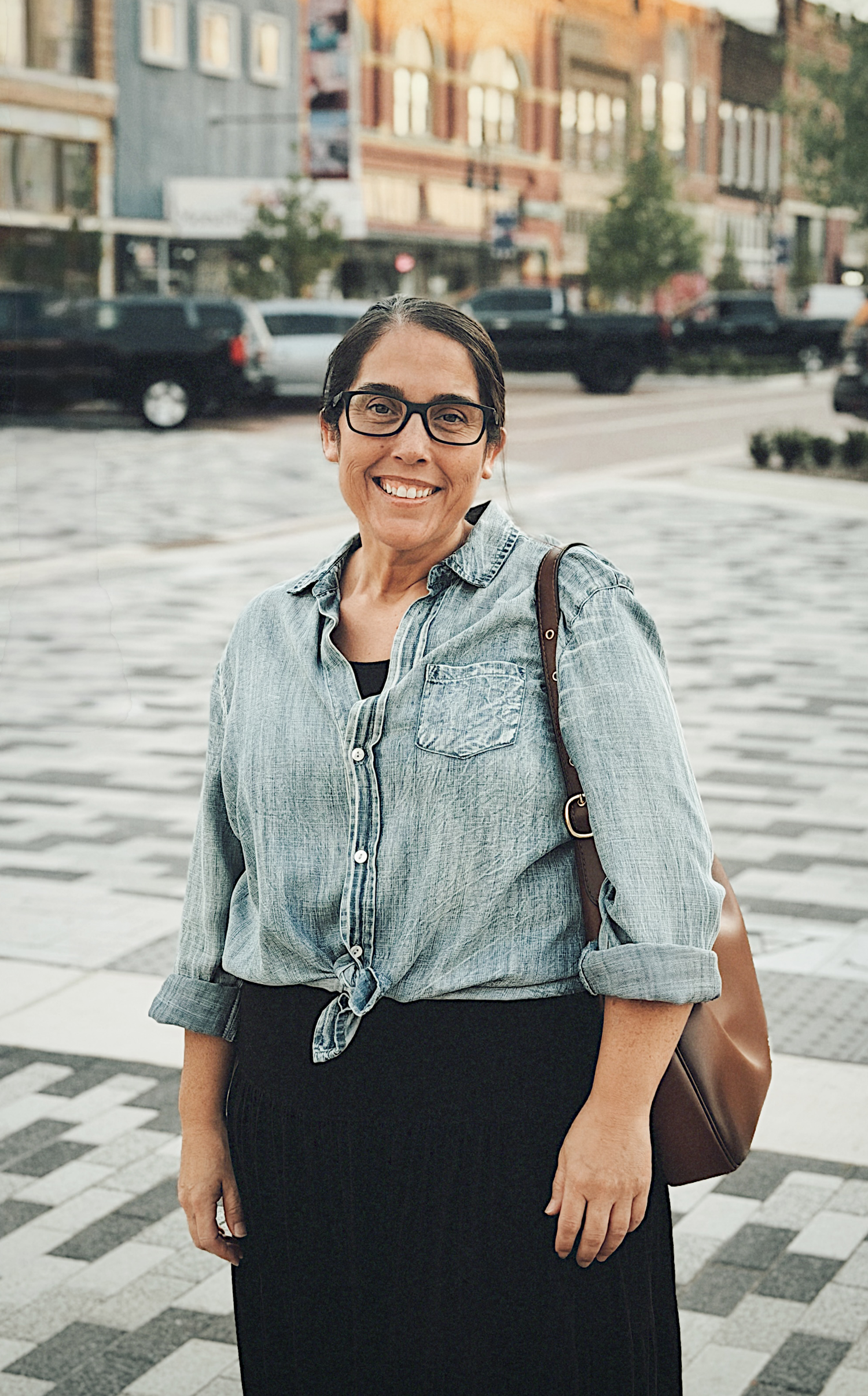 Smiling woman in denim shirt and black skirt stands in urban plaza with shops in background.