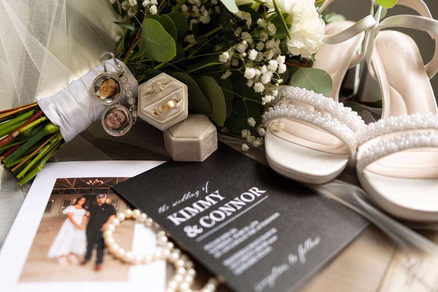 Wedding flat lay featuring invitation for Kimmy and Connor, bouquet, ring box, heels, and photo charms.