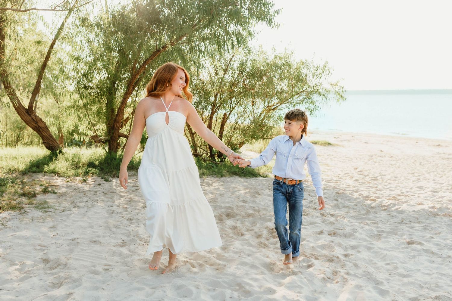 Mother in white dress holding hands with young son walking on sandy beach near water.