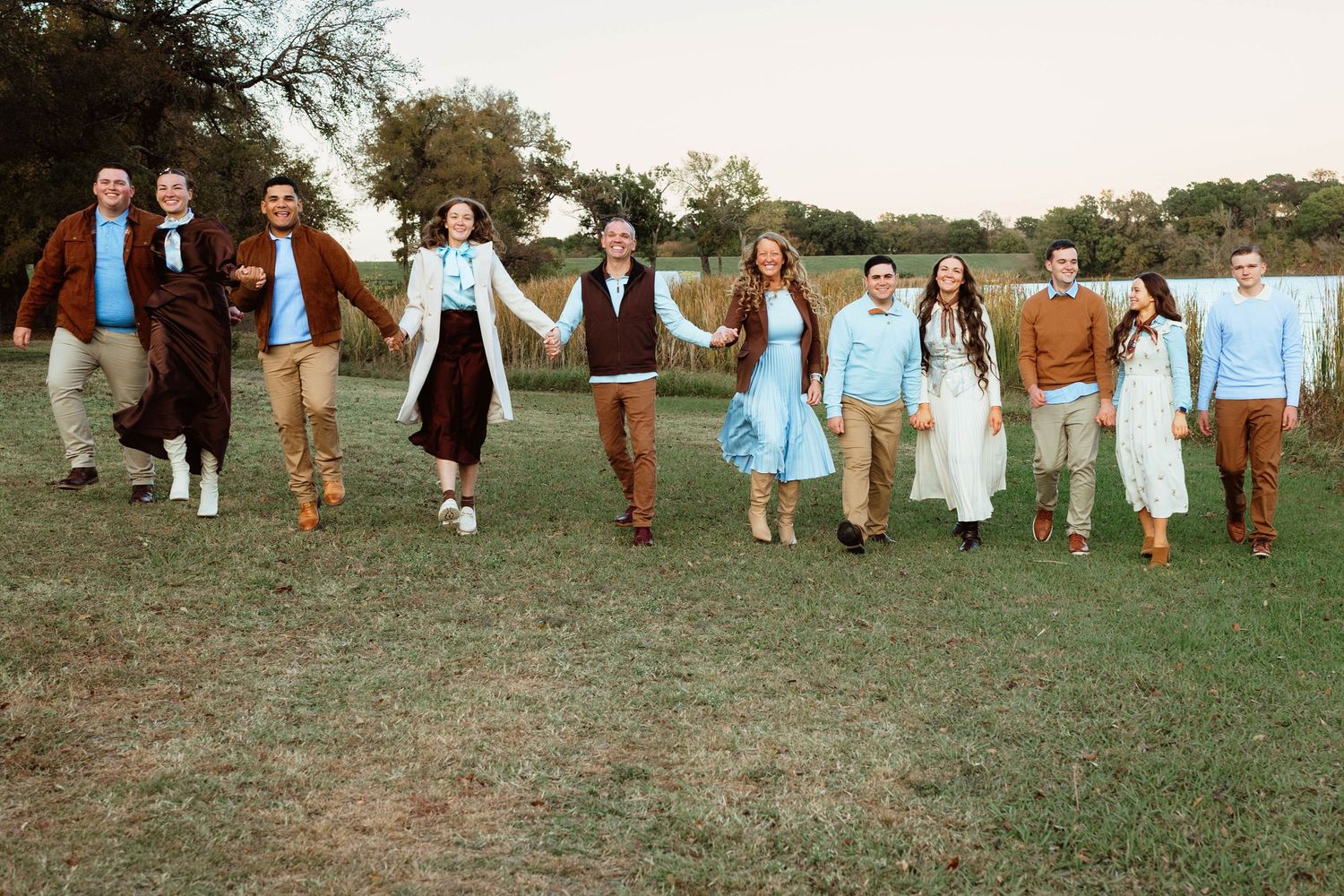 Large group of people hold hands and walk together across a green field near a lake outdoors.