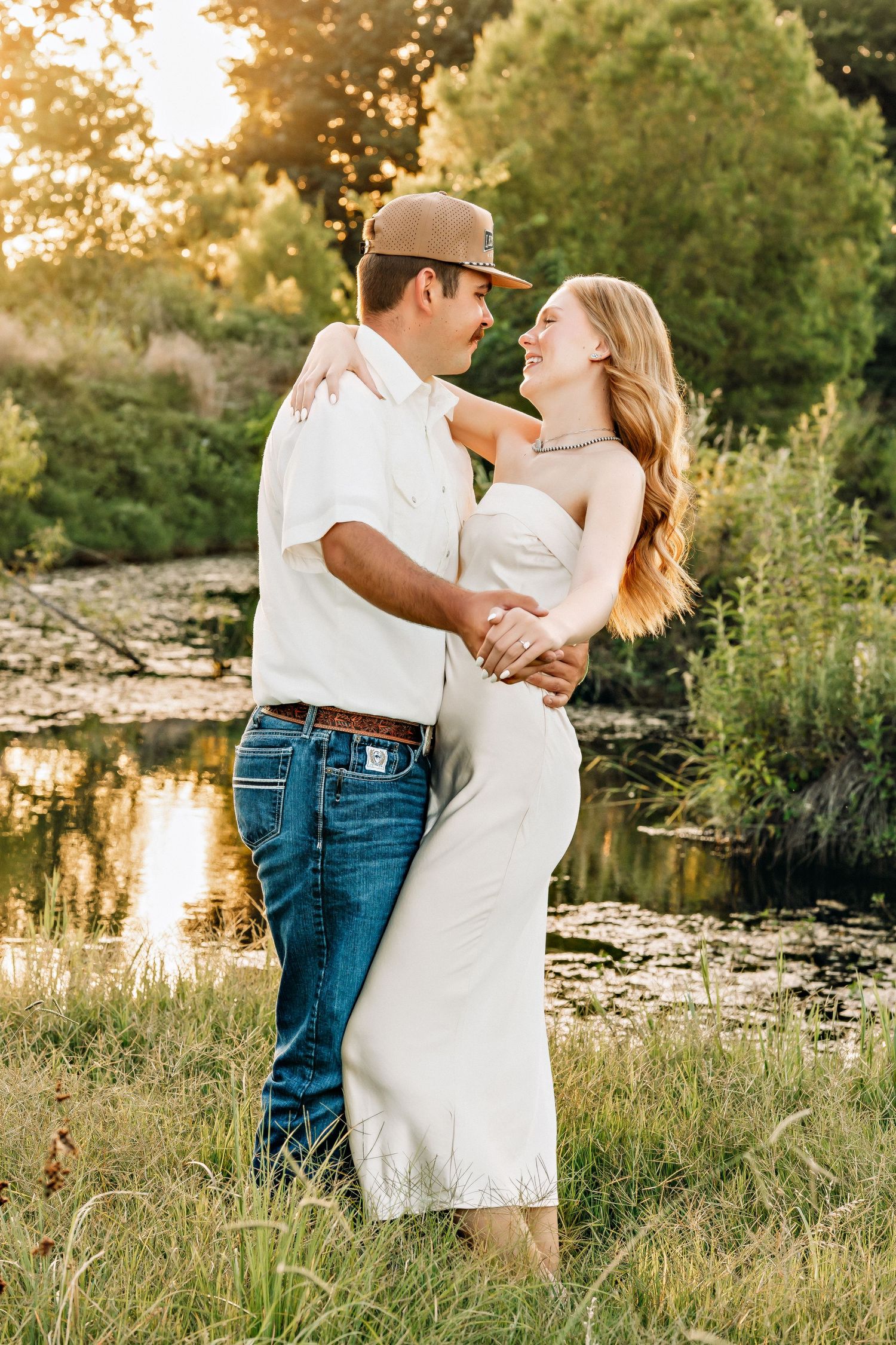 A couple embraces and gazes at each other near a reflective pond surrounded by lush greenery at sunset.