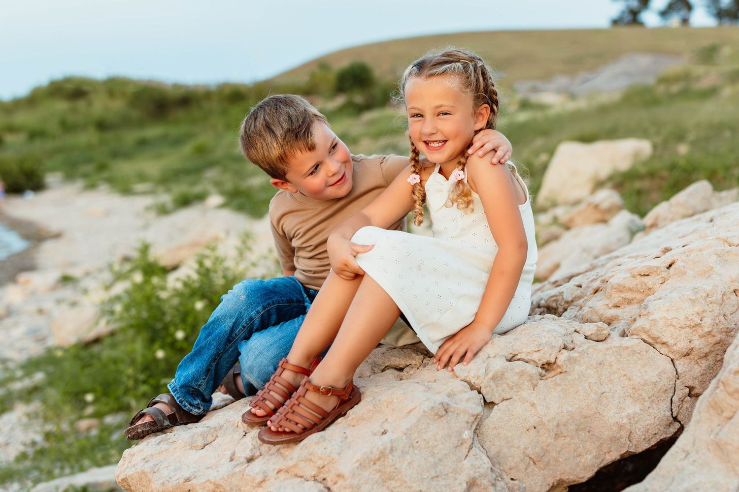 Two young children sitting on rocks outdoors, smiling together in a candid sibling portrait photo.