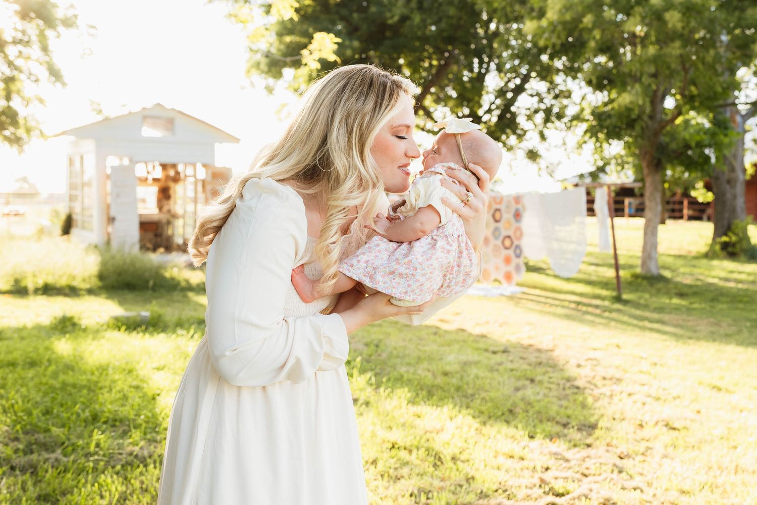 A blonde woman in a white dress kisses her newborn baby while standing in a sunny green backyard.