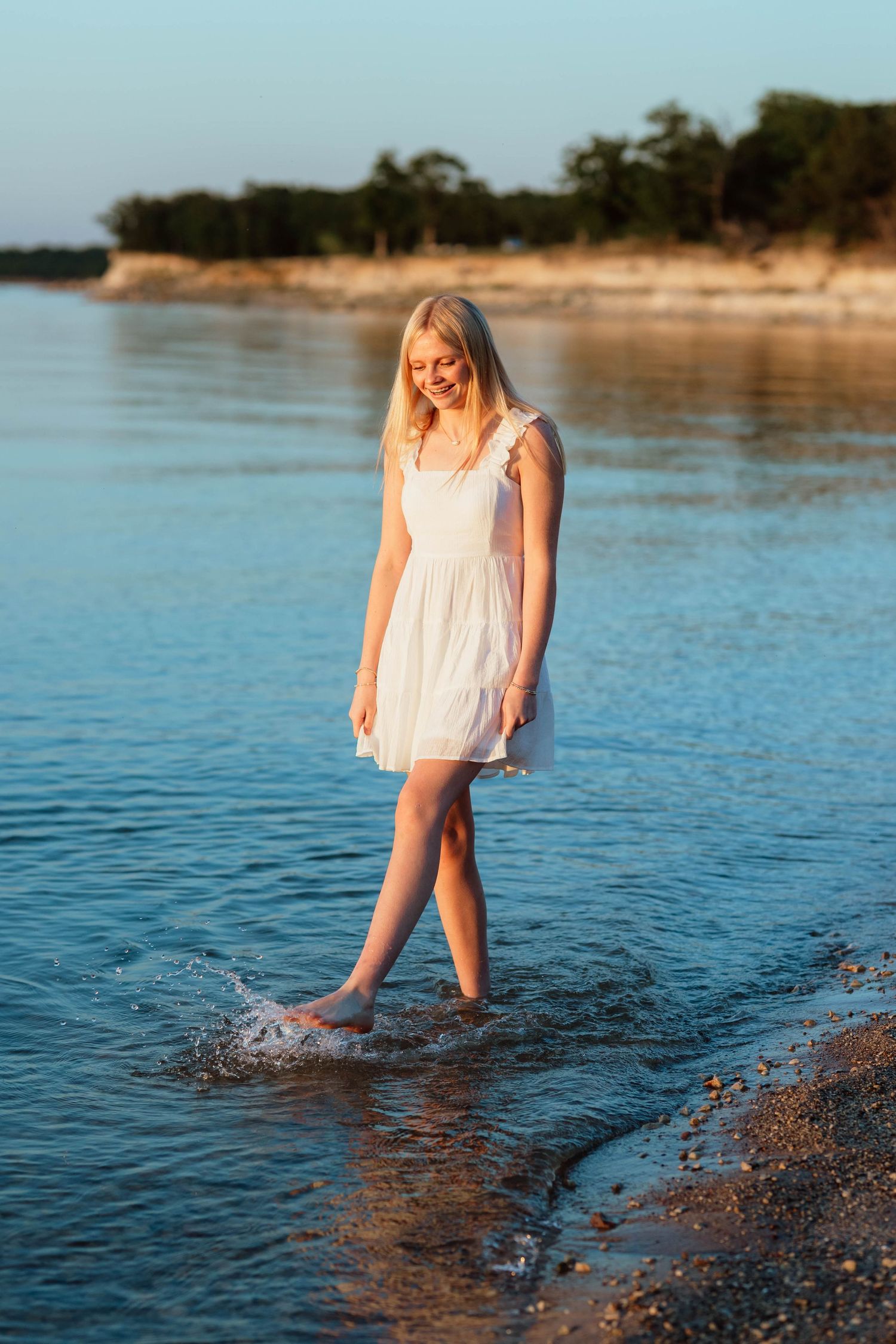 Smiling blonde woman in white dress wading in calm lake water at golden hour.