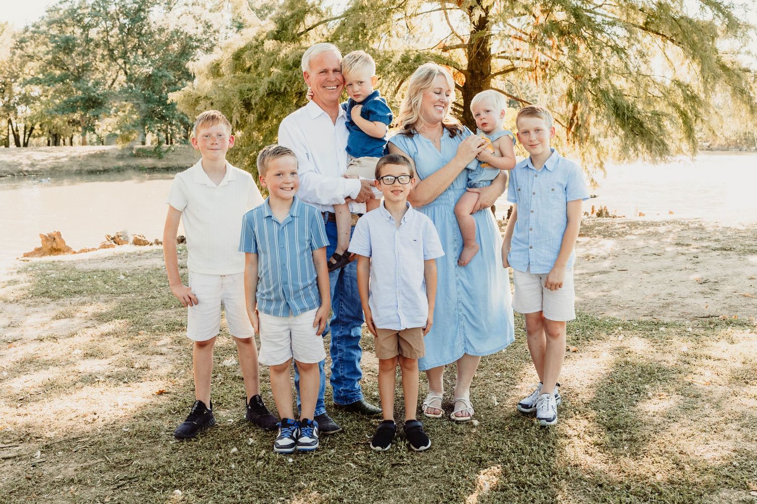Large family portrait with grandparents and five boys smiling outdoors under a tree in coordinating blue and white outfits.