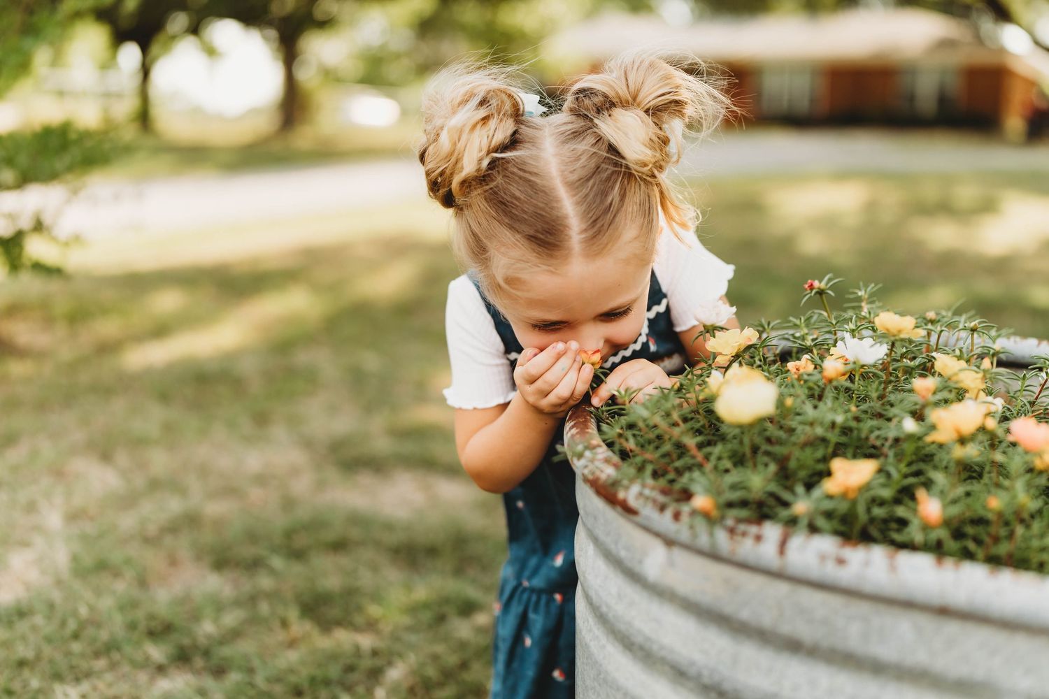 A blonde toddler girl smells flowers in a vintage galvanized metal tub planter in a garden setting.