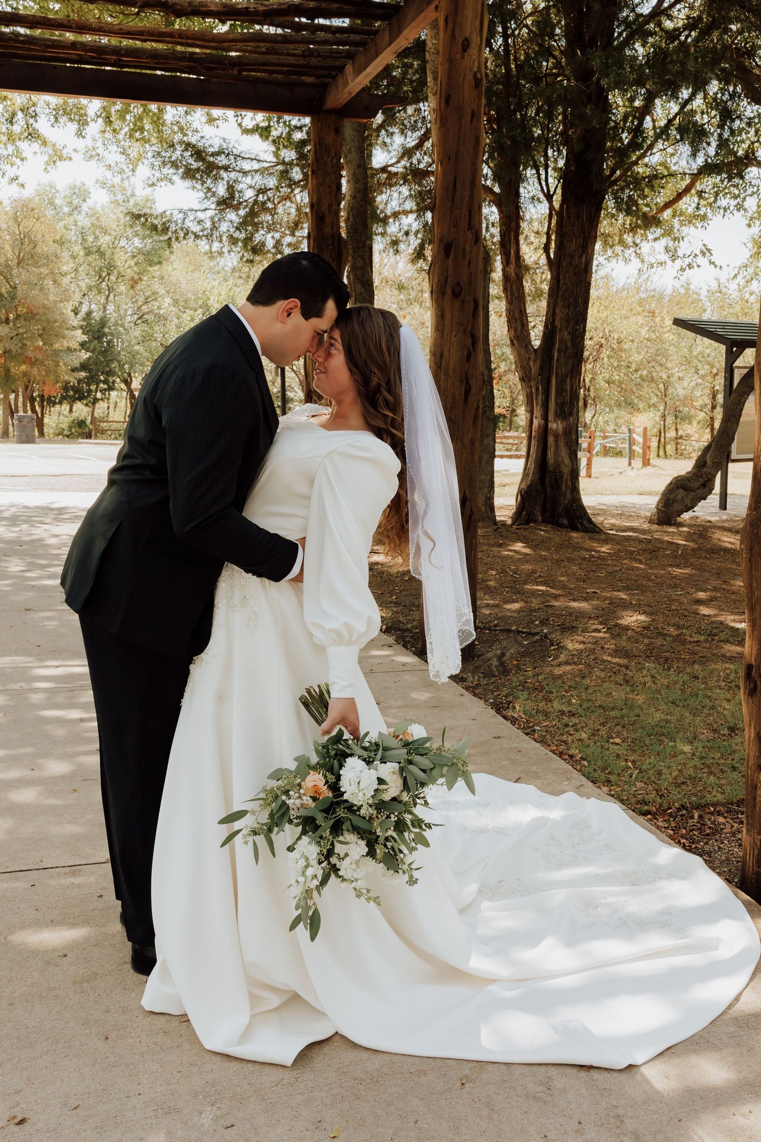Bride in off-shoulder white gown and groom in black suit kissing under outdoor pergola.
