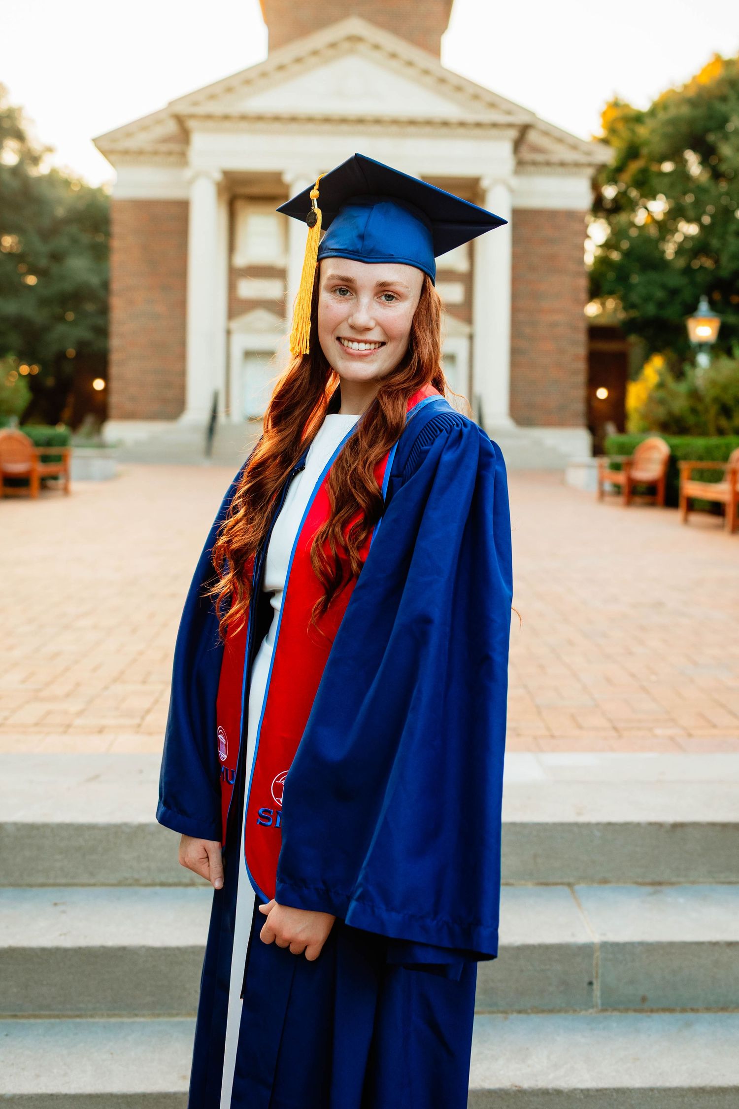 Smiling redhead woman in blue graduation cap and gown on university steps.