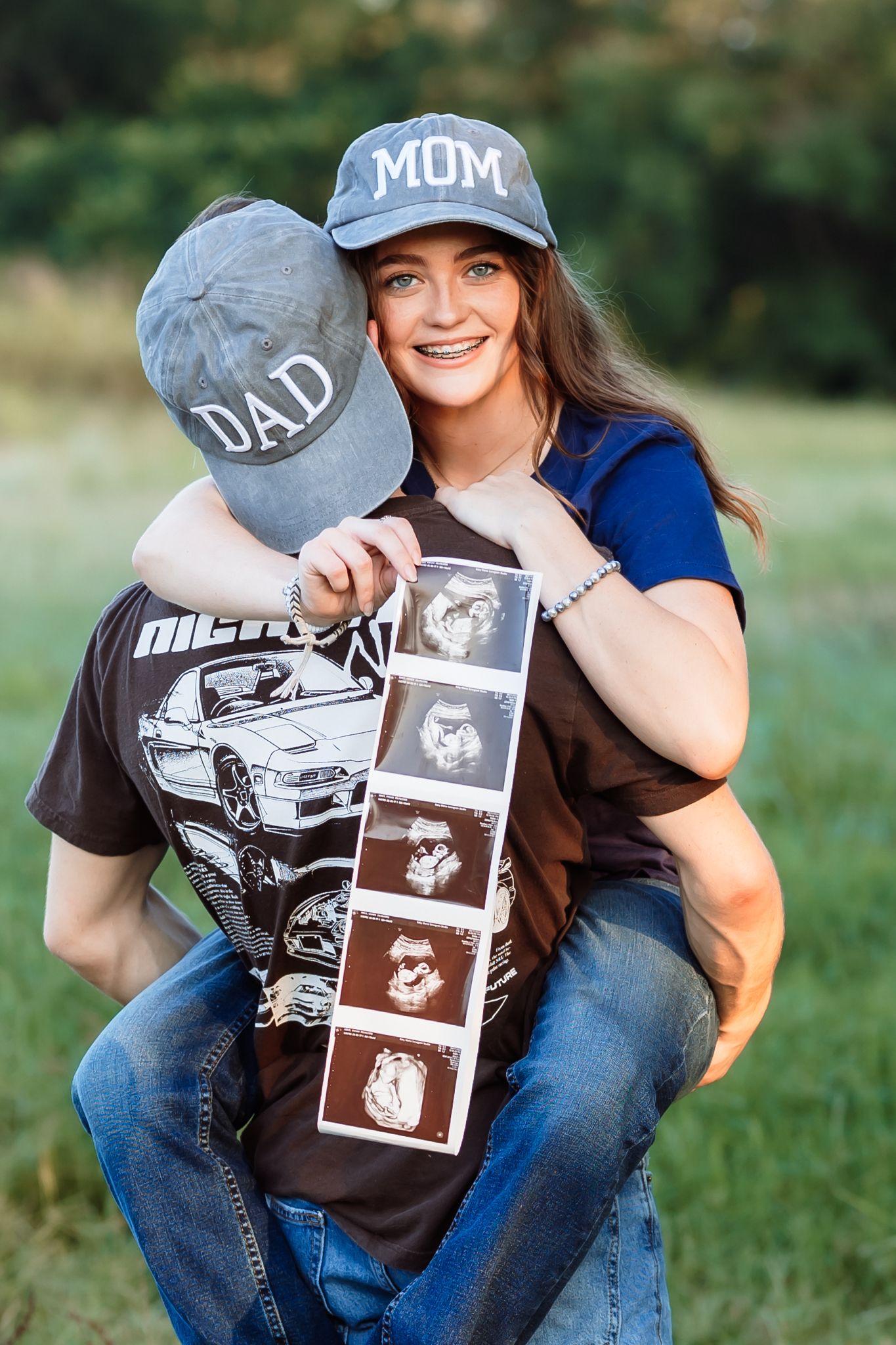 Smiling woman wearing a Mom hat holds ultrasound photos while man wearing a Dad hat carries her outdoors.