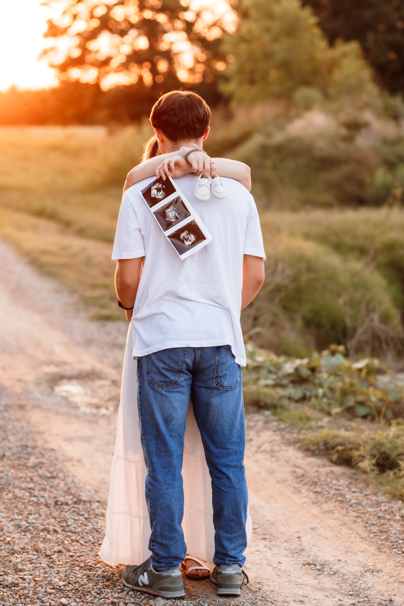 Couple from behind at sunset holding ultrasound photos and tiny baby shoes on a rural dirt road.