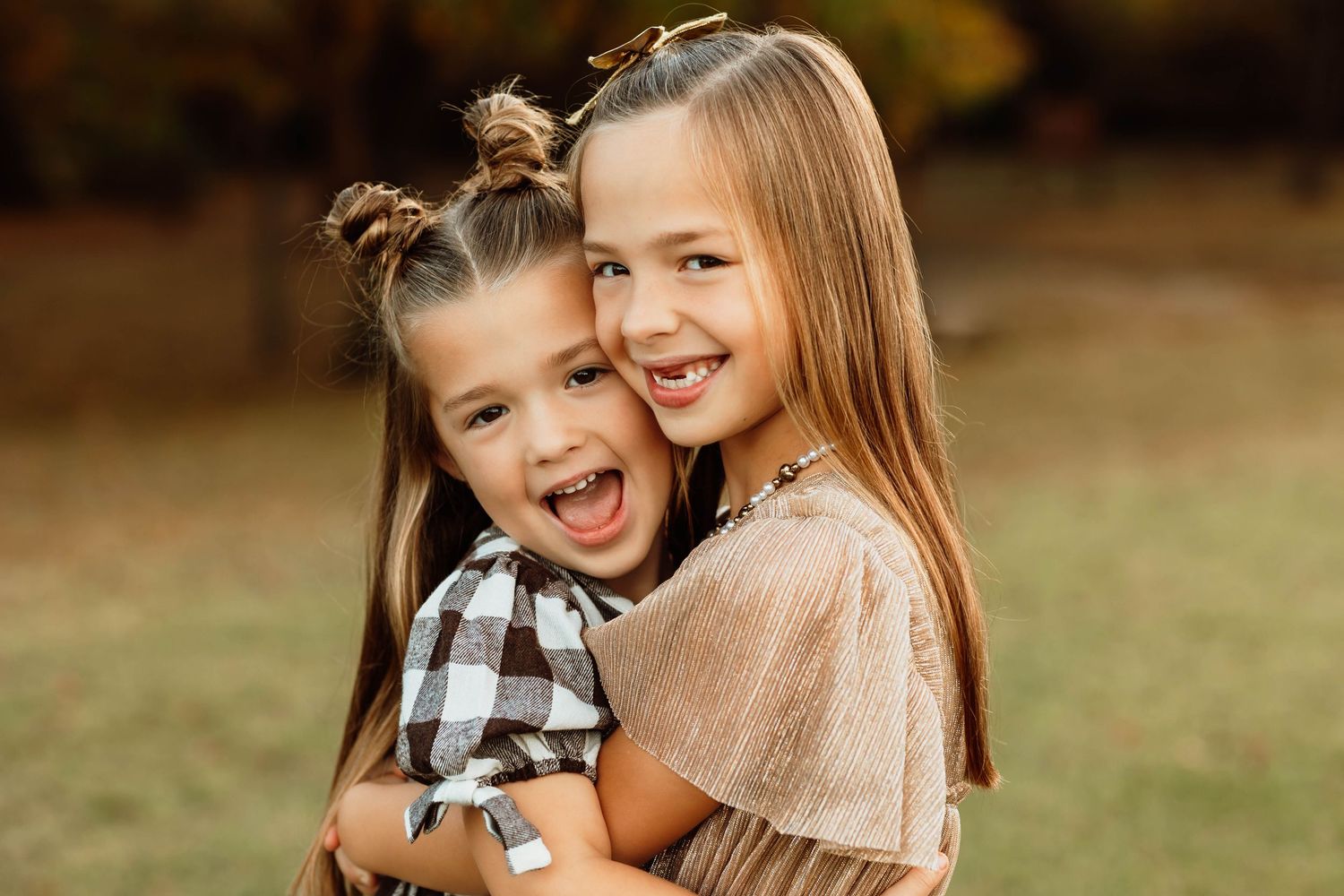 Two young smiling sisters hug each other outdoors with autumn foliage in the background.