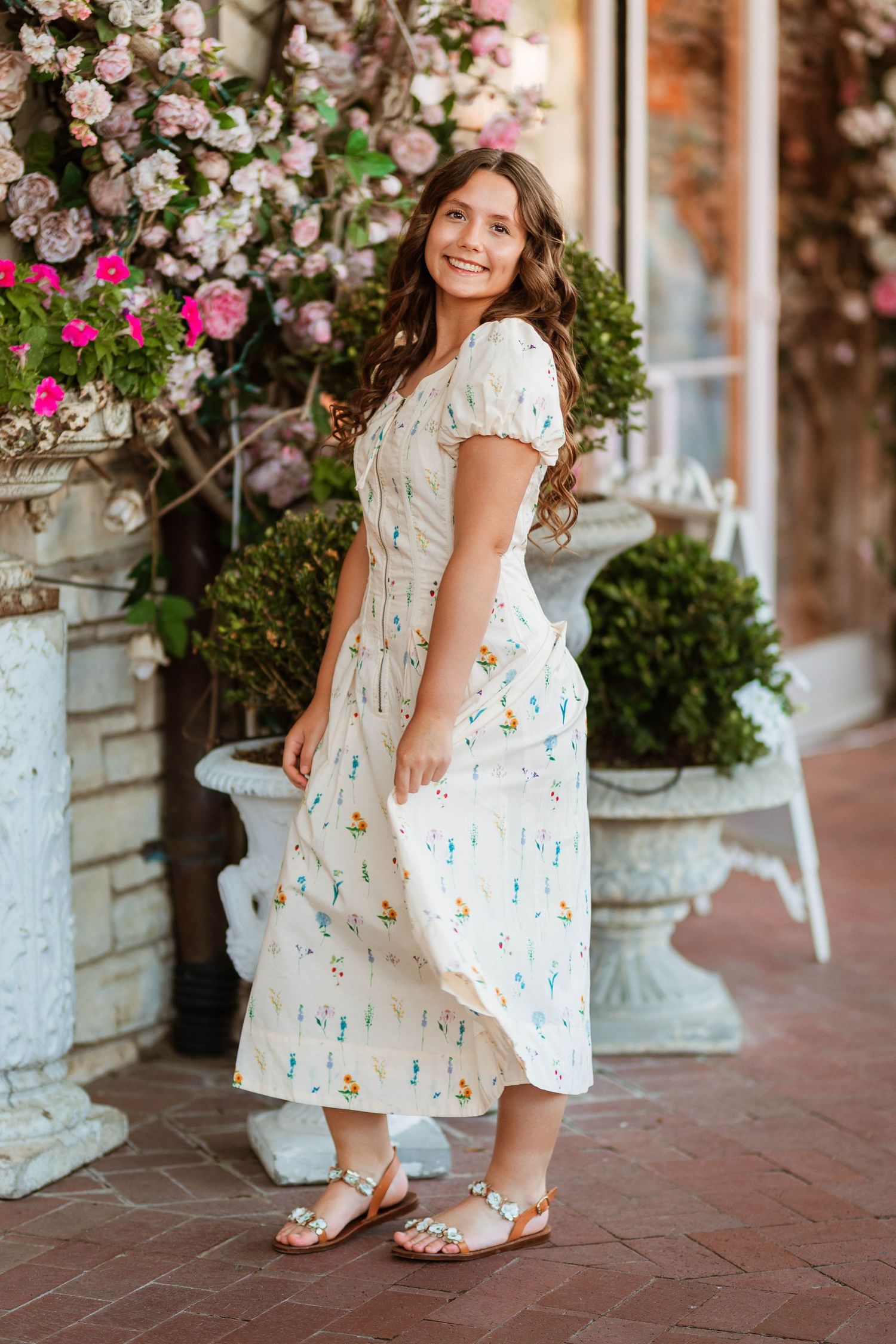 Smiling brunette woman in floral midi dress posing beside pink flower display.