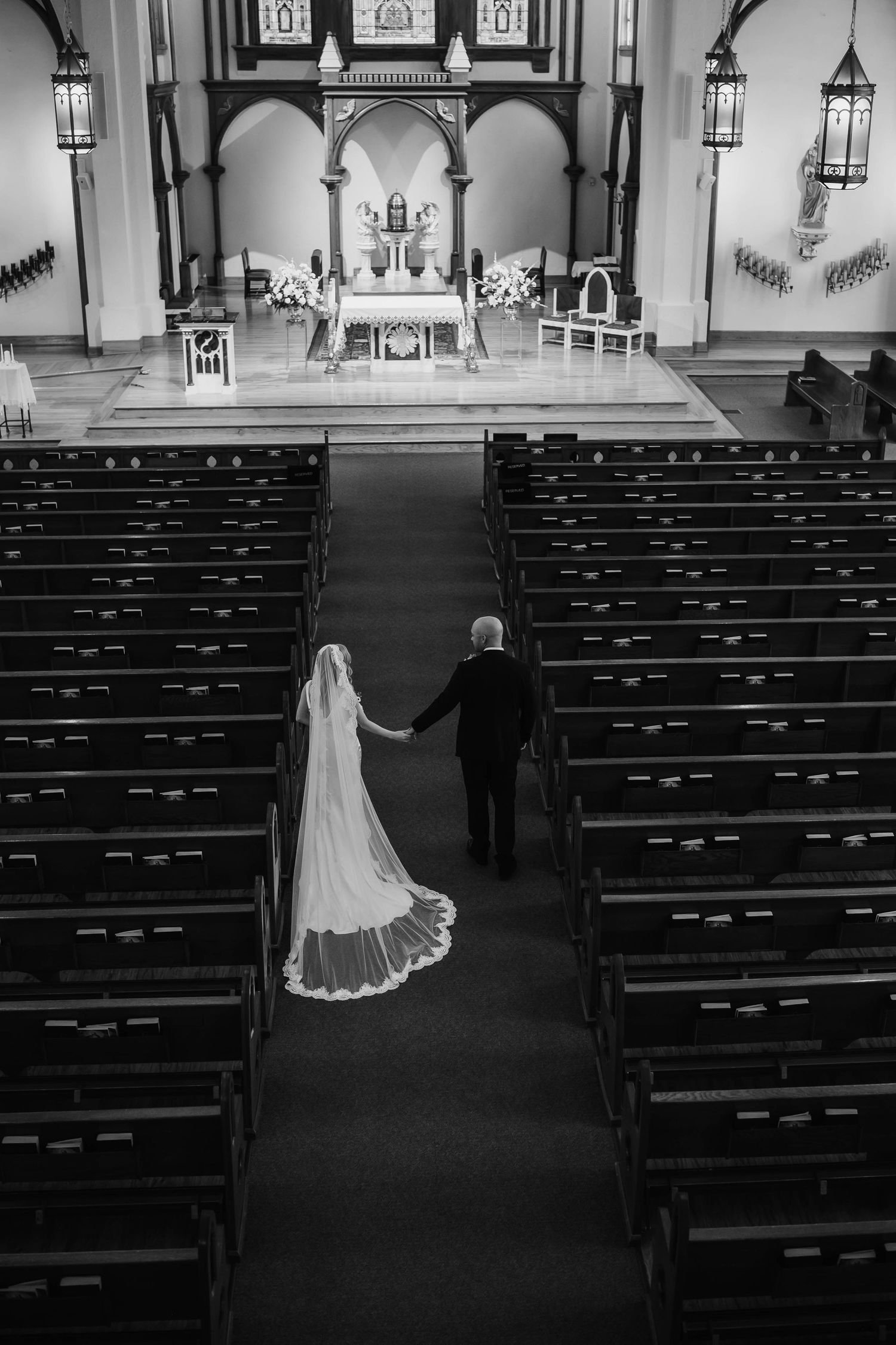 Black and white overhead view of bride and groom walking toward church altar holding hands.