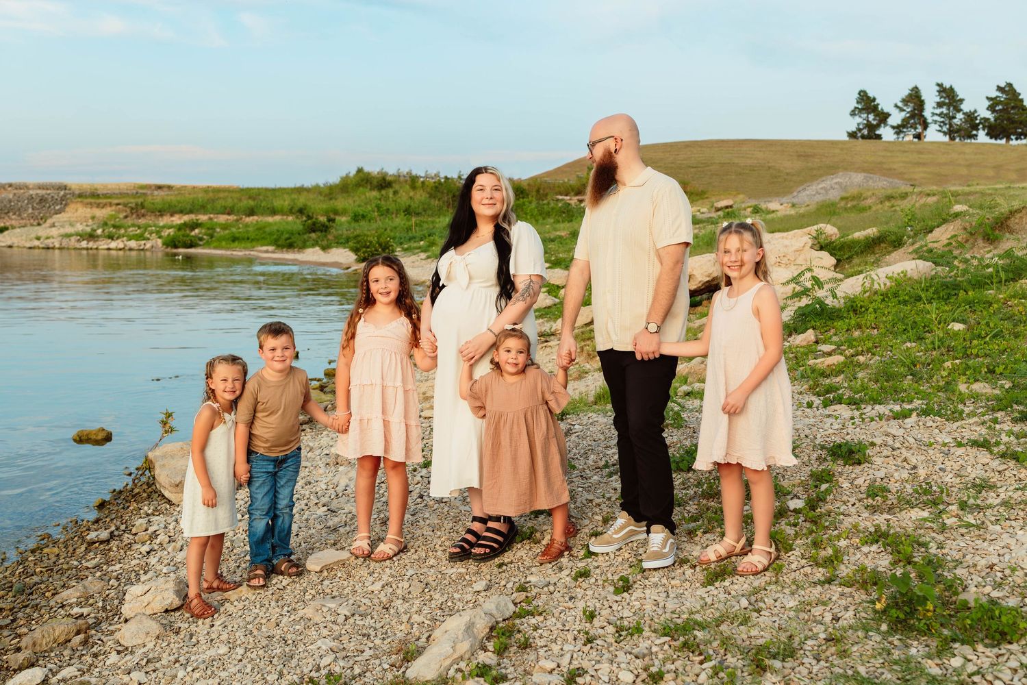 Large family of seven poses together near a scenic river with rolling green hills in the background.