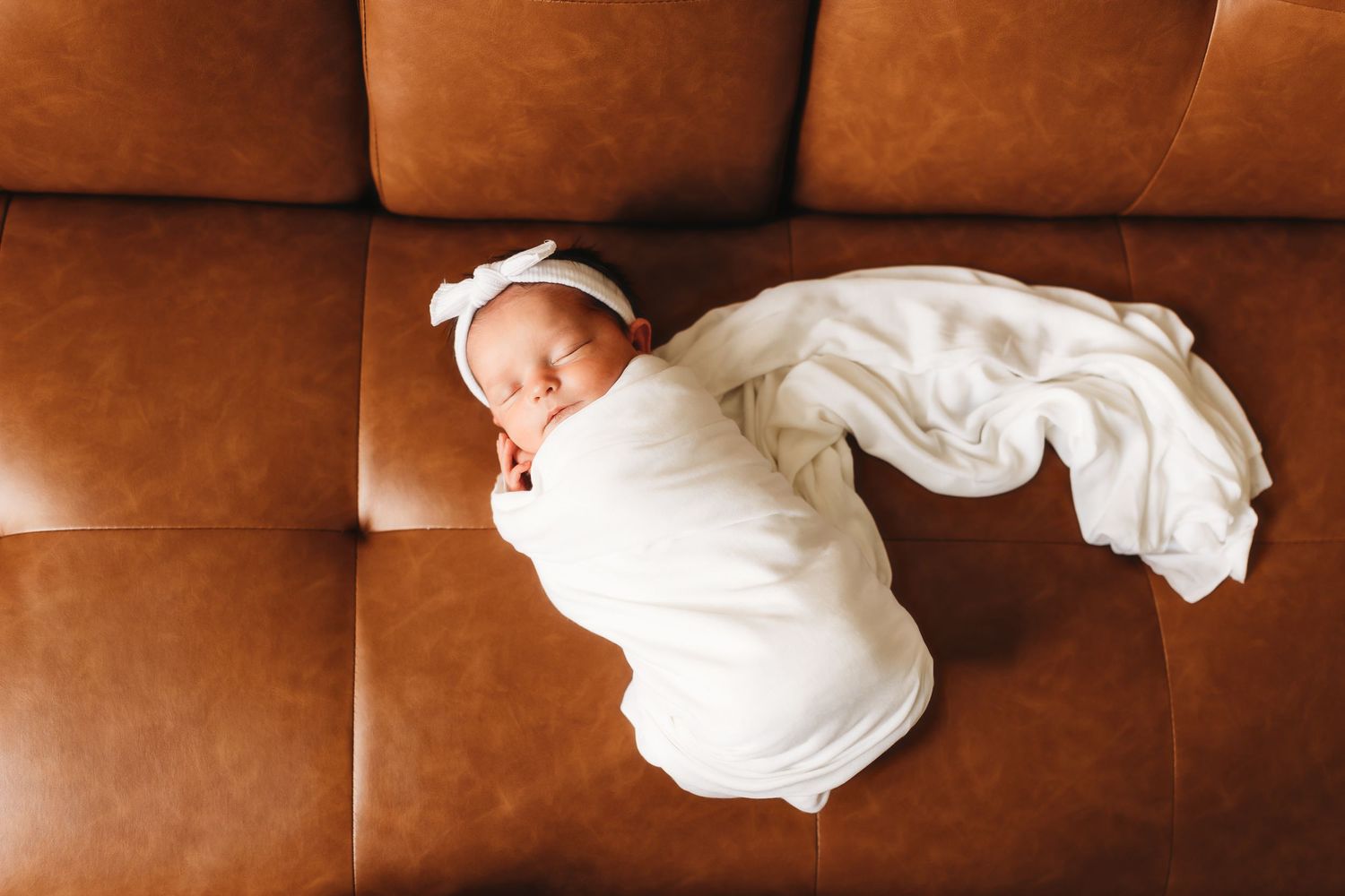 Sleeping newborn baby wrapped in white swaddle with bow headband lying on brown leather couch.