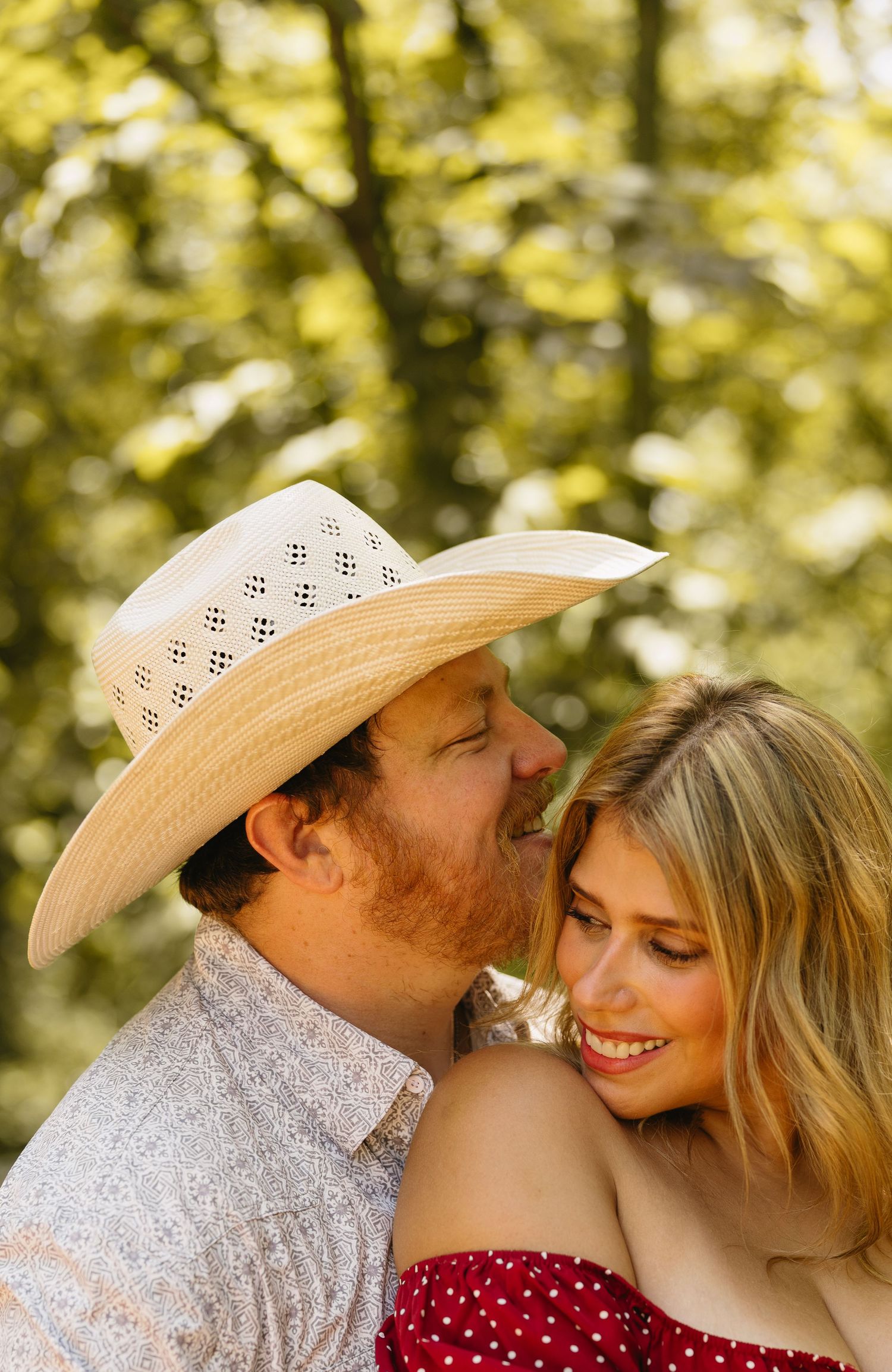 A smiling couple outdoors, man in straw cowboy hat kissing woman in red polka dot top with green trees behind.