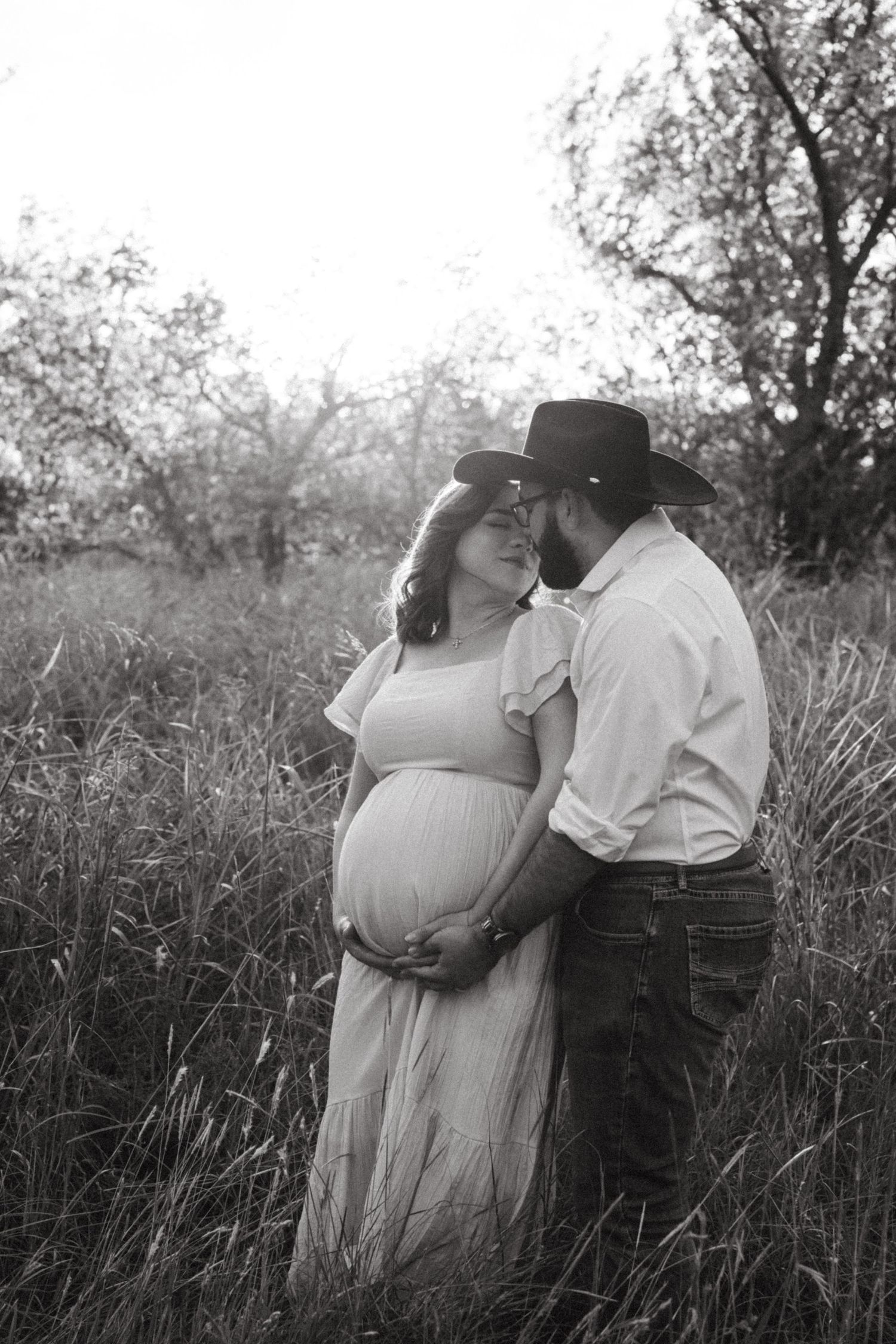 Black and white photo of couple embracing in field during maternity shoot.