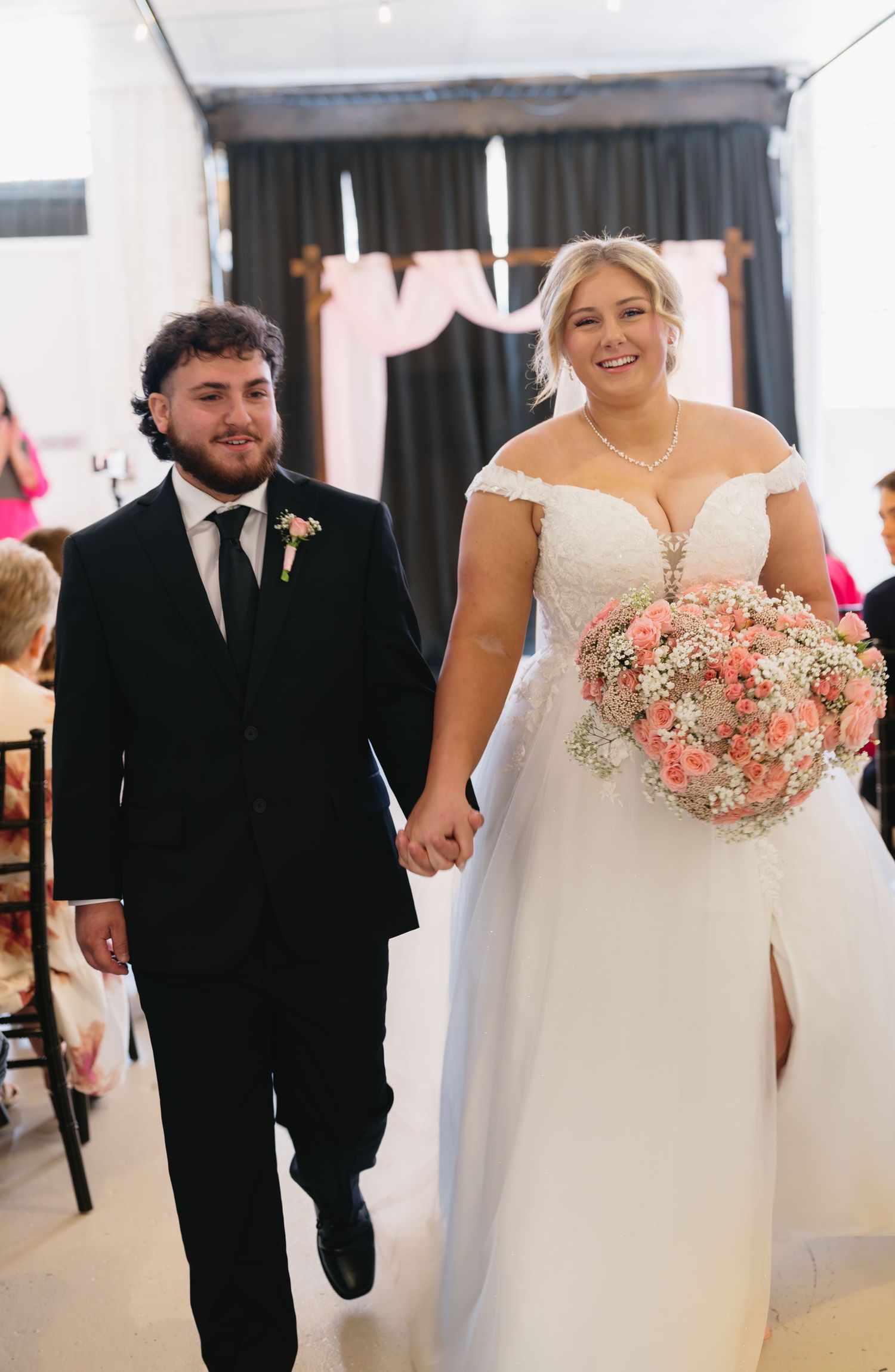 Smiling bride in white off-shoulder gown and groom in black suit walk hand-in-hand down wedding aisle.