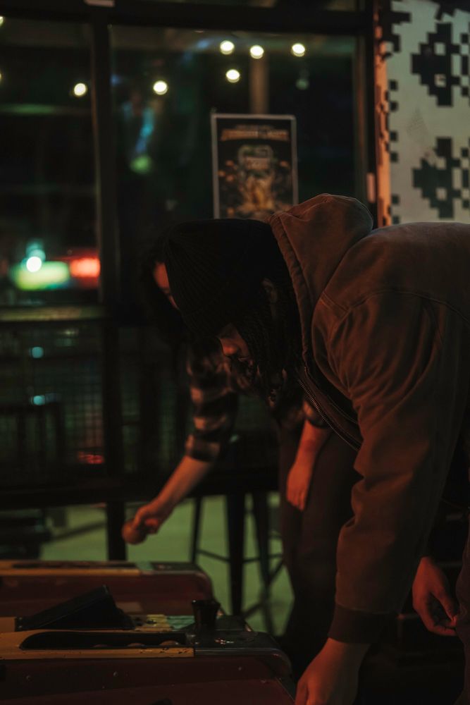 Person in hooded jacket leaning over arcade pinball machine in dimly lit neon-lit game room at night.