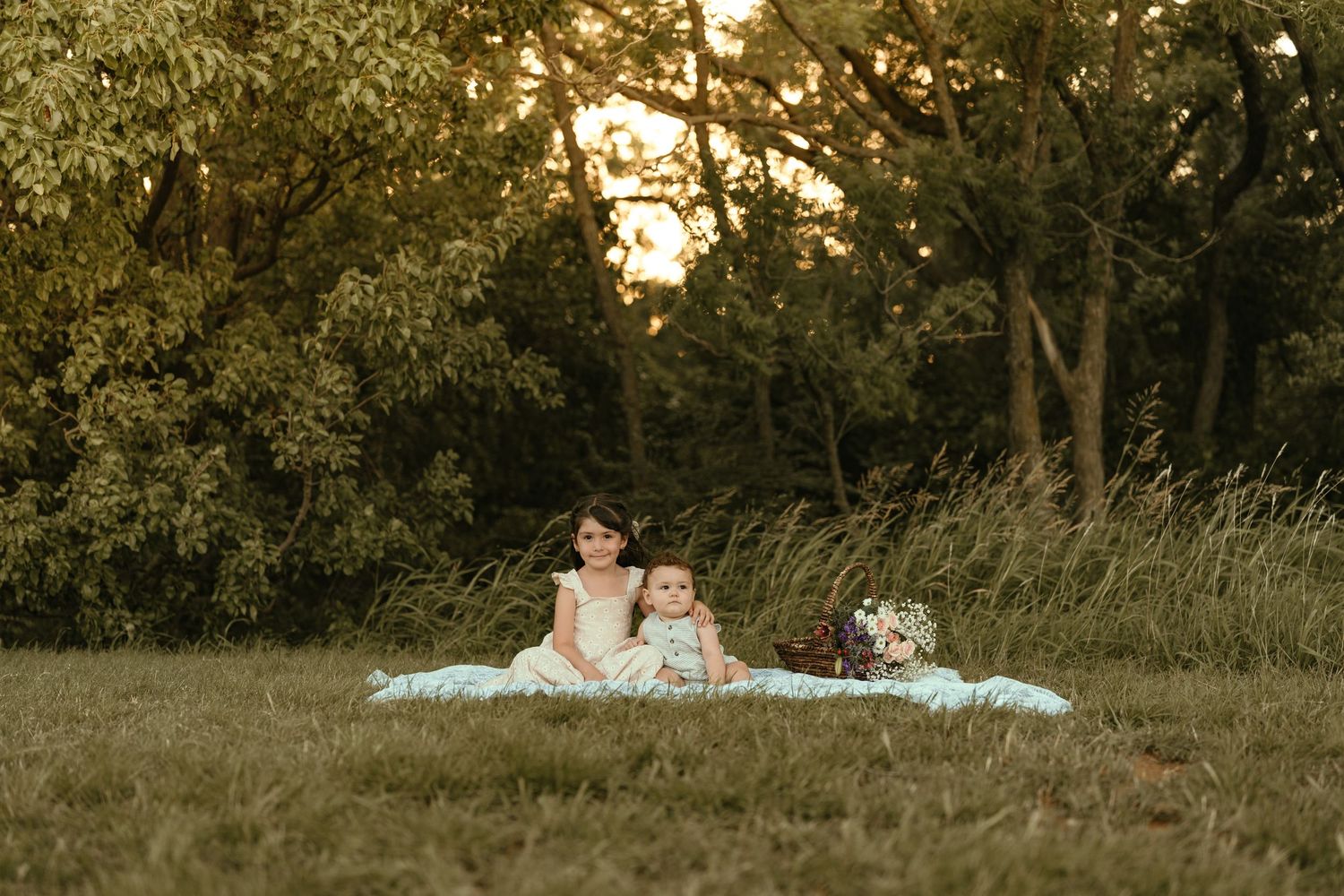 Two young children sit together on a white blanket in a grassy field surrounded by trees at sunset.