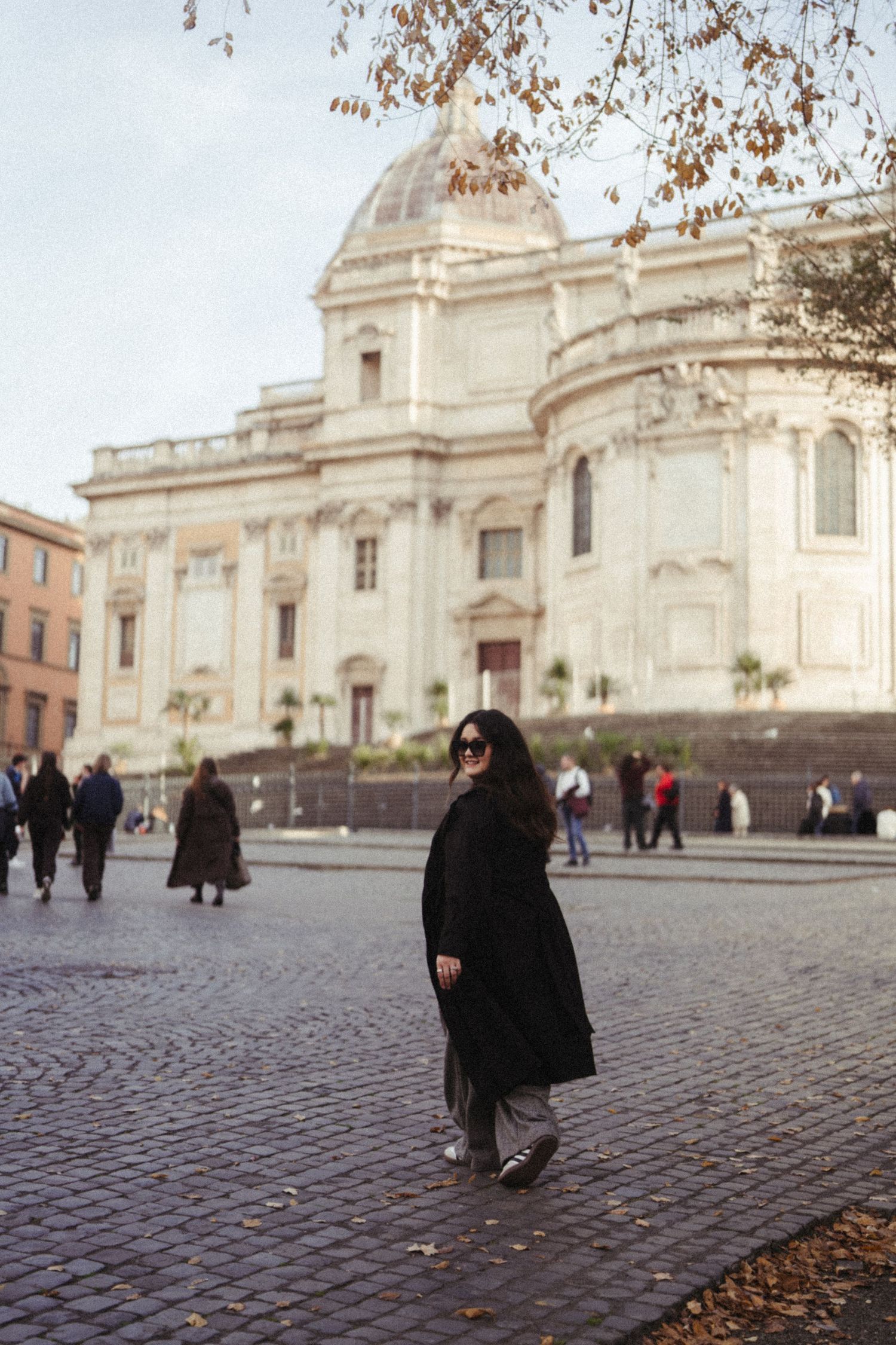 Woman in black coat posing in front of a grand historic building on cobblestone plaza in Rome.