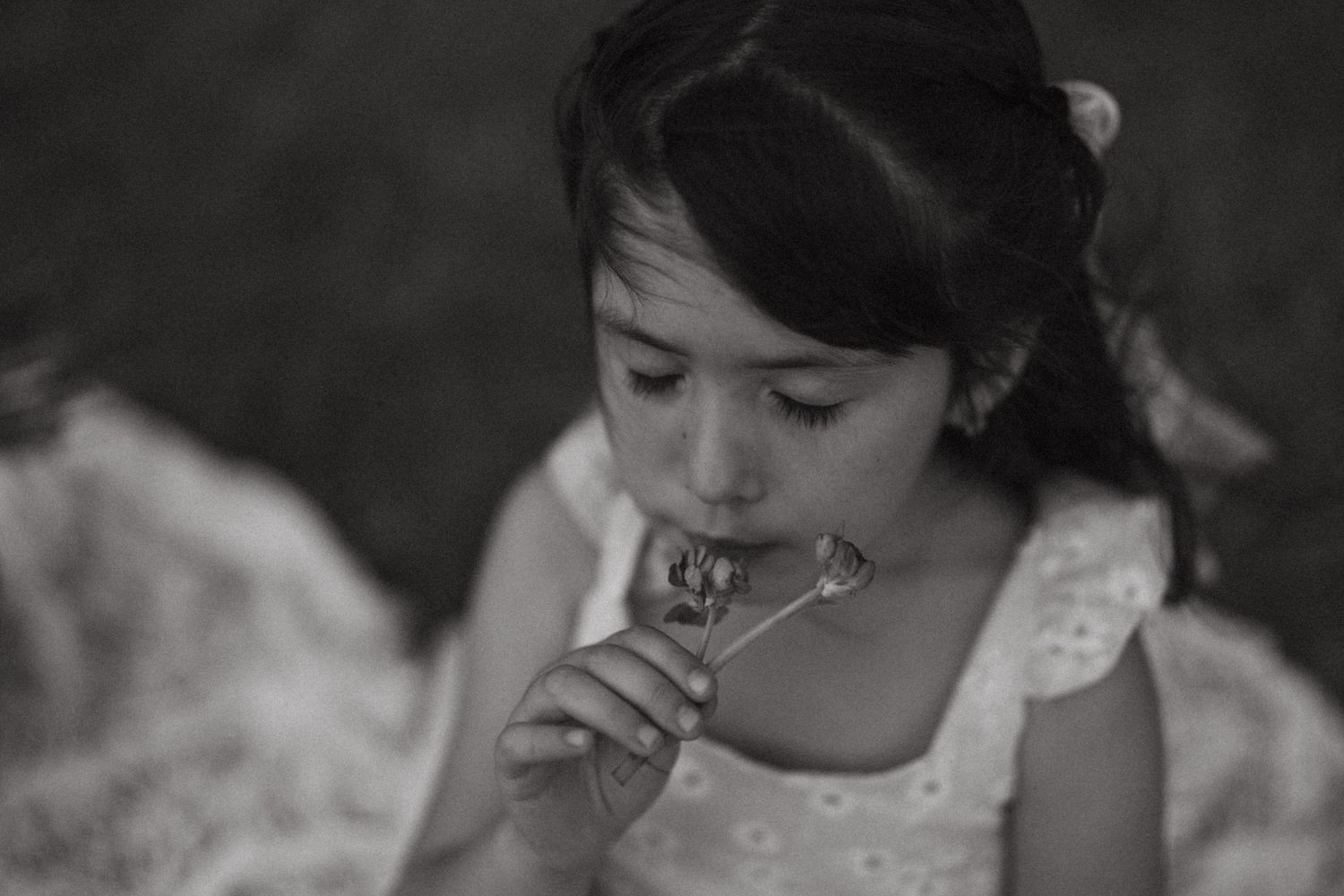 Black and white close-up of a young girl in a white dress smelling a small flower.