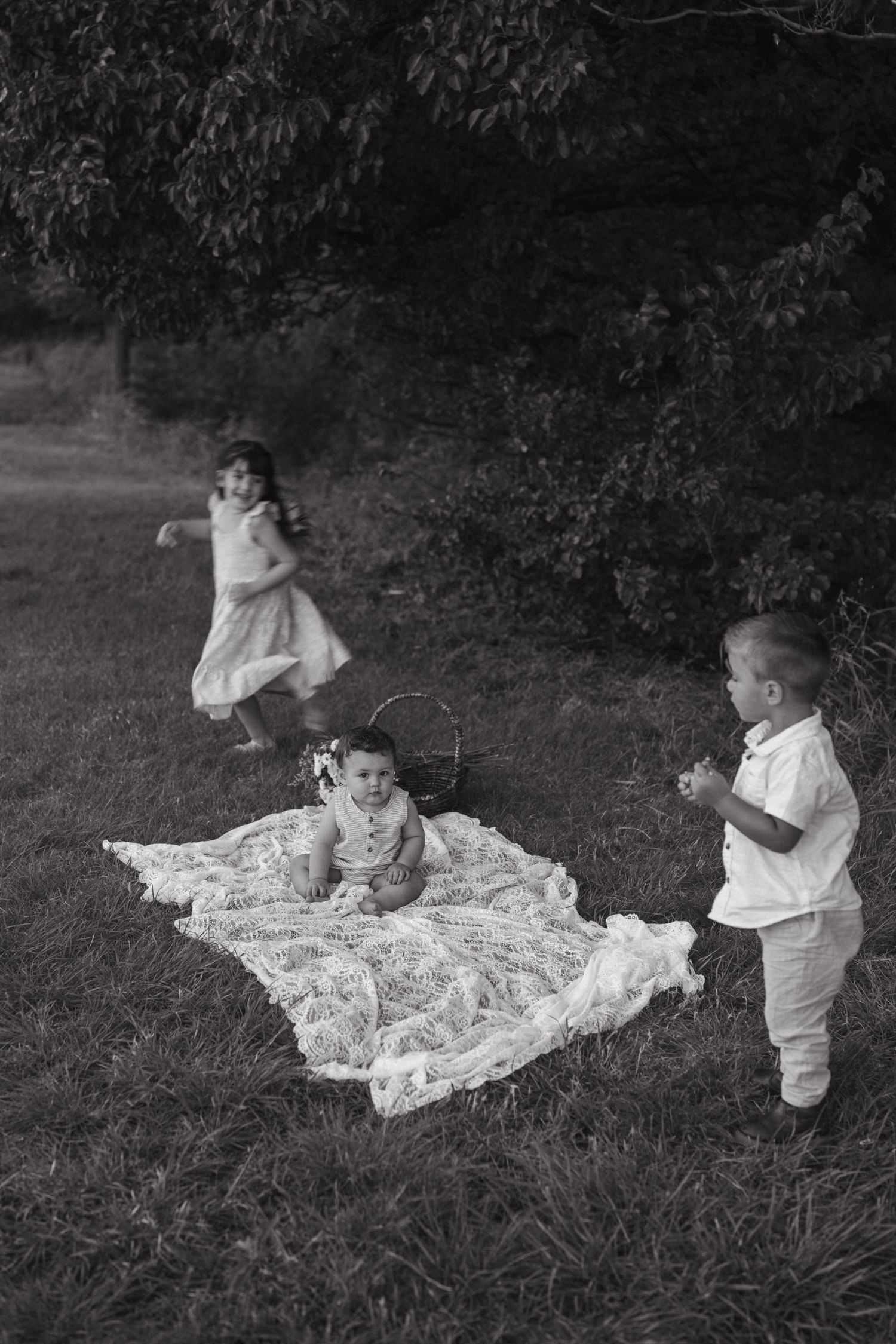 Black and white photo of three children playing on a lace blanket under trees in a yard.