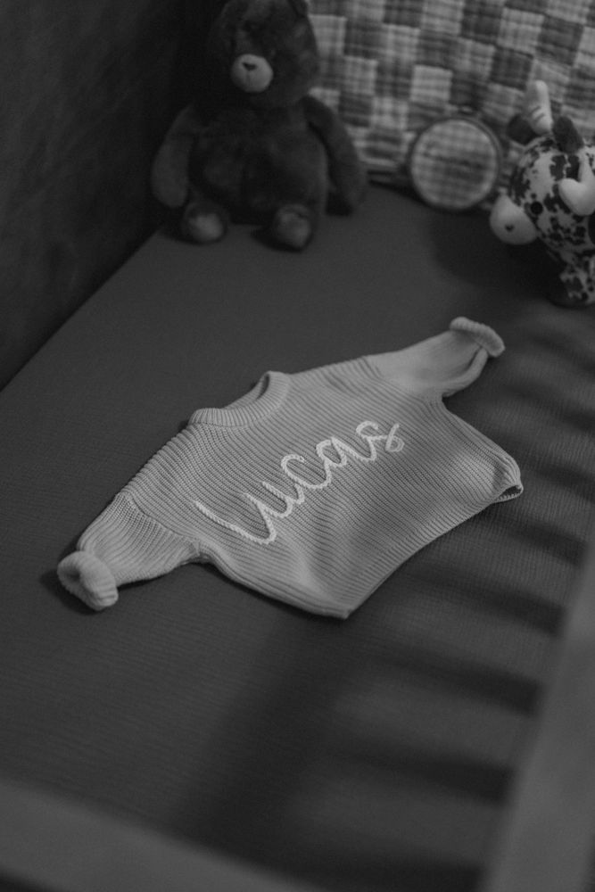 Black and white photo of a personalized knit baby sweater reading Lucas in a crib with toys.