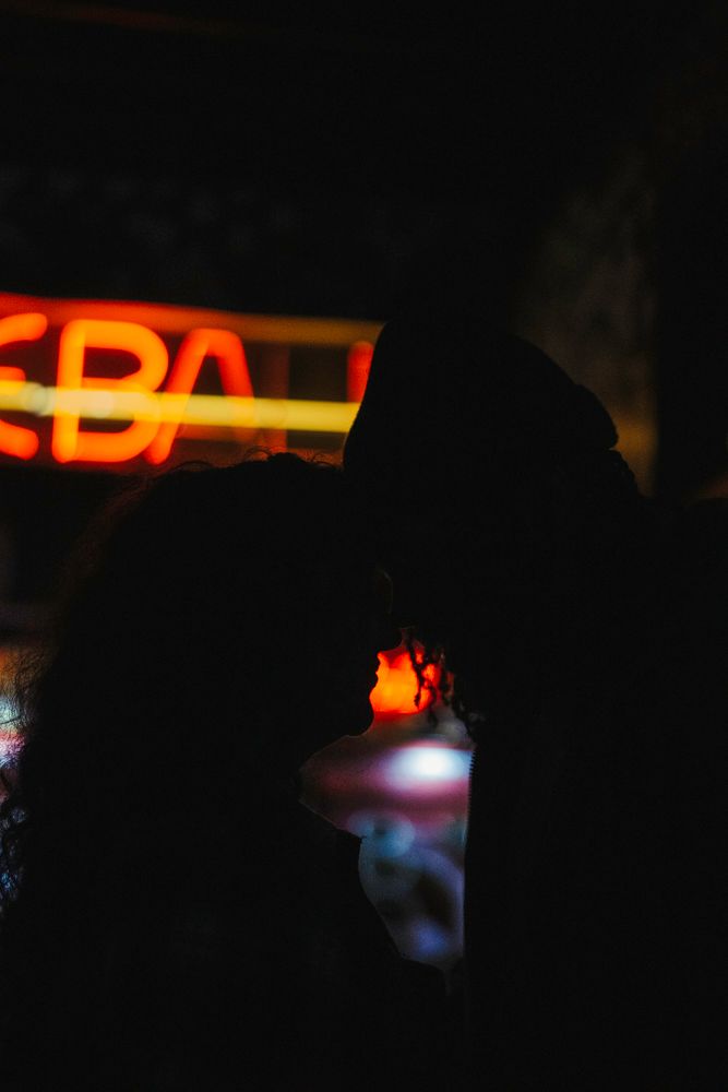 Romantic silhouette of couple about to kiss in front of glowing red and orange neon bar sign at night.