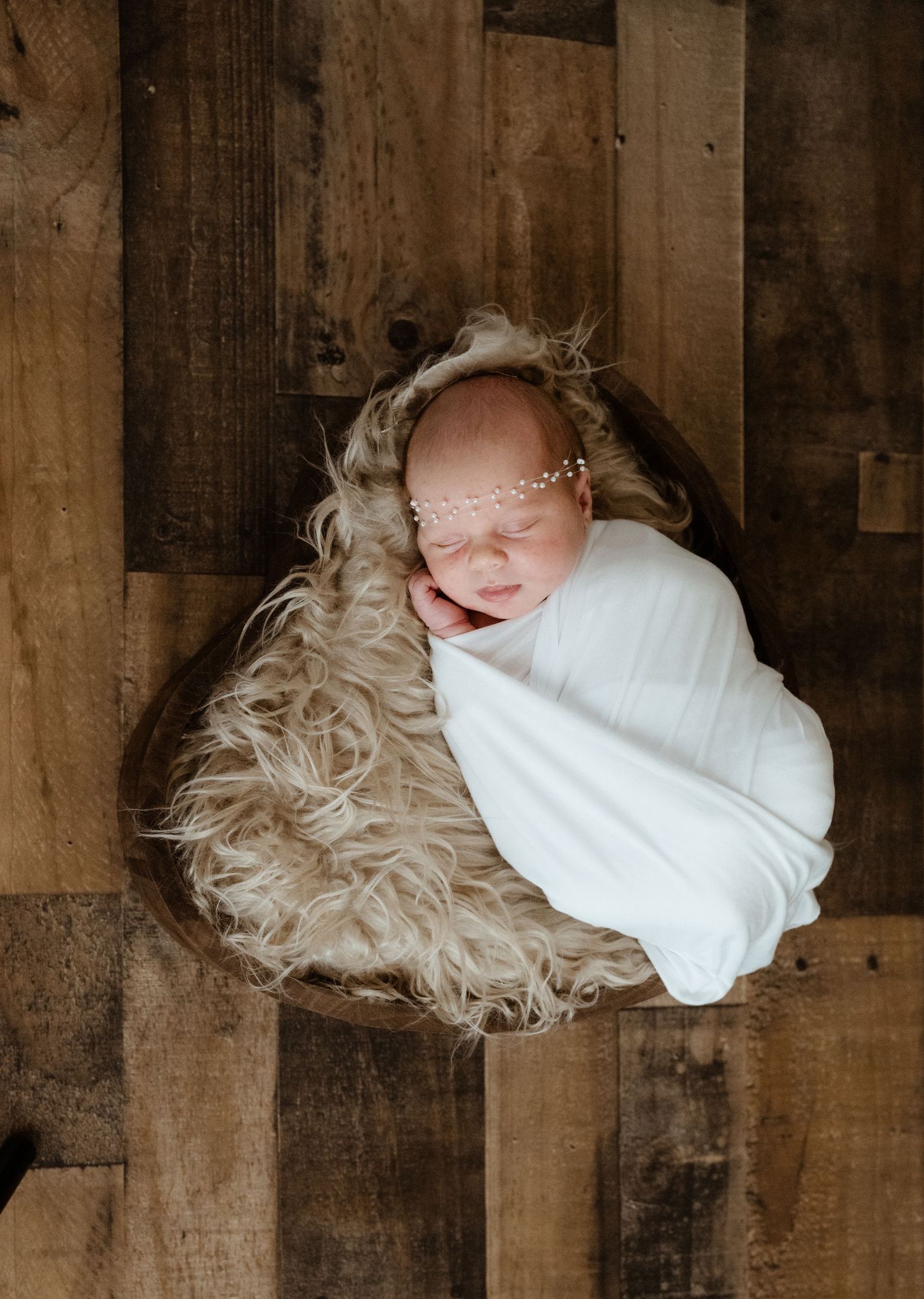 Sleeping newborn baby swaddled in white wrap with floral headband, nestled in fluffy bowl on rustic wood background.