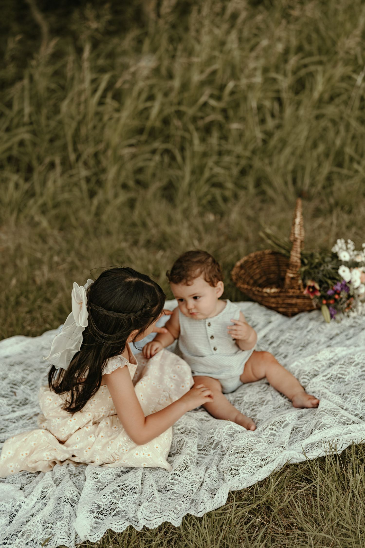 A mother in lace dress tenderly interacts with her baby on a white blanket in a lush green field.