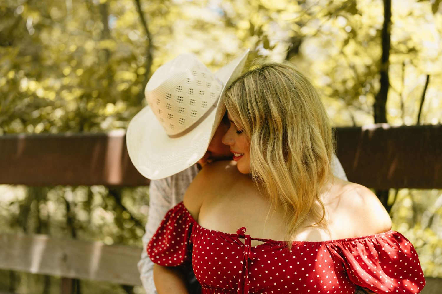 Two blonde women in a sunny outdoor setting, one wearing a white cowboy hat and the other a red polka dot off-shoulder top.