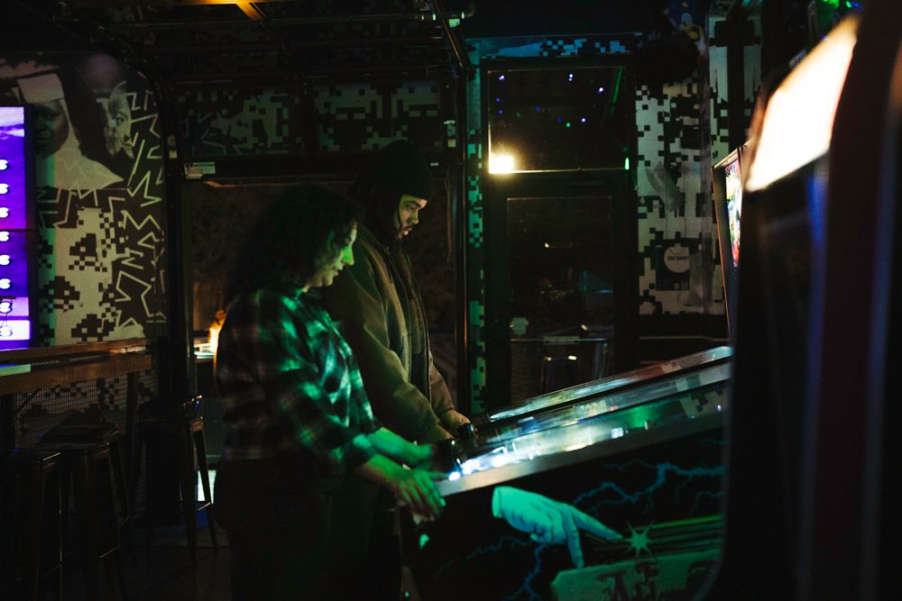 Couple playing pinball machine together in dark neon-lit arcade bar with colorful green ambient lighting.