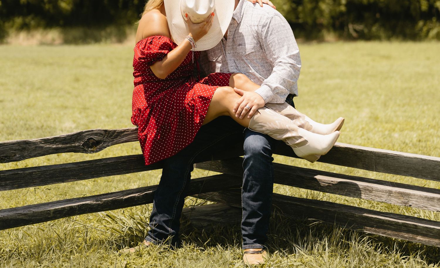 Couple in western attire sitting on a wooden fence, woman in red polka dot dress and man in white cowboy hat.