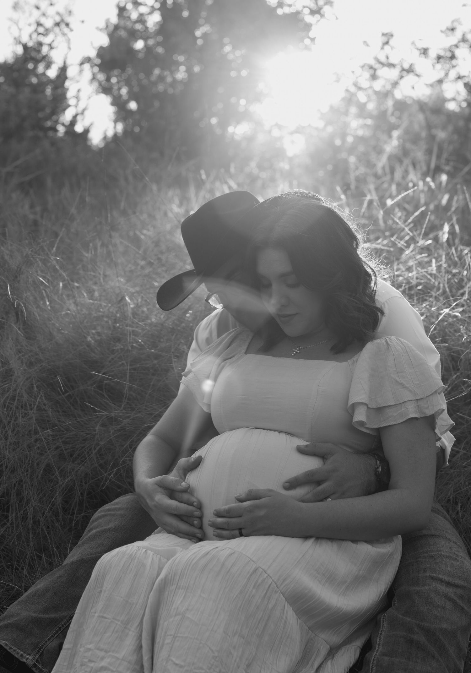 Black and white maternity photo of couple embracing outdoors in golden sunlight, man in cowboy hat cradling pregnant belly.