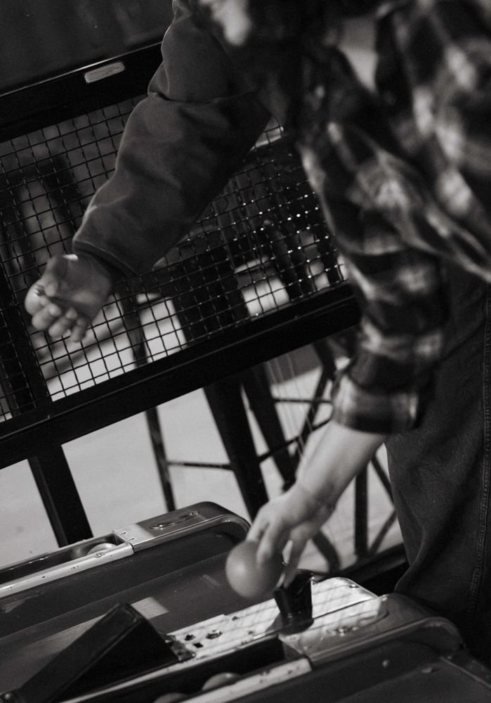 Black and white close-up of two people playing pinball machine, hands reaching across the game surface.