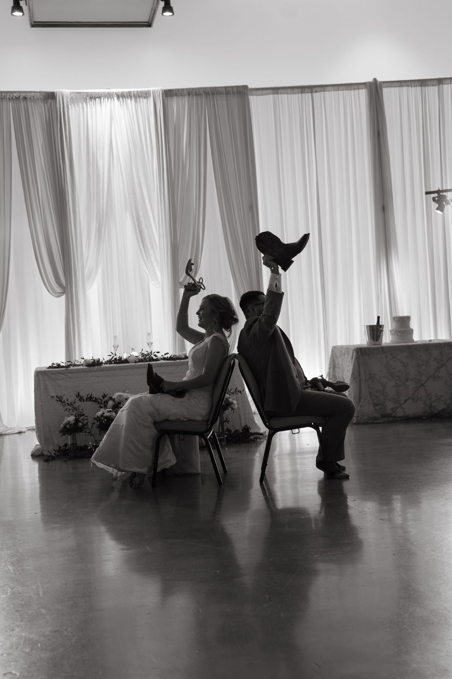 Black and white silhouette of bride and groom seated in chairs raising shoes during fun wedding game.