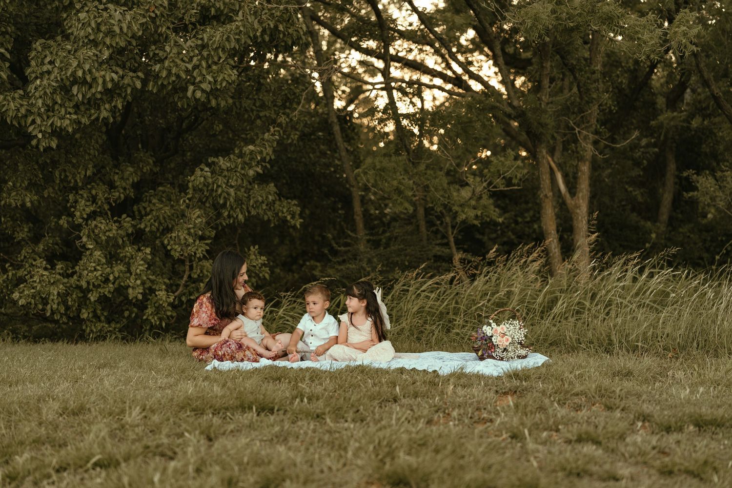 A family with young children relaxes together on a white blanket in a lush green meadow at golden hour.