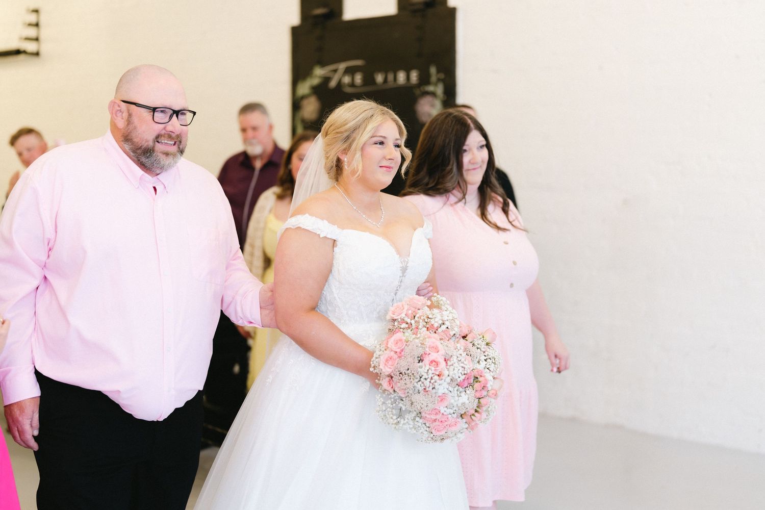 Bride in white gown with pink bouquet walks down aisle escorted by man in pink shirt at indoor wedding.